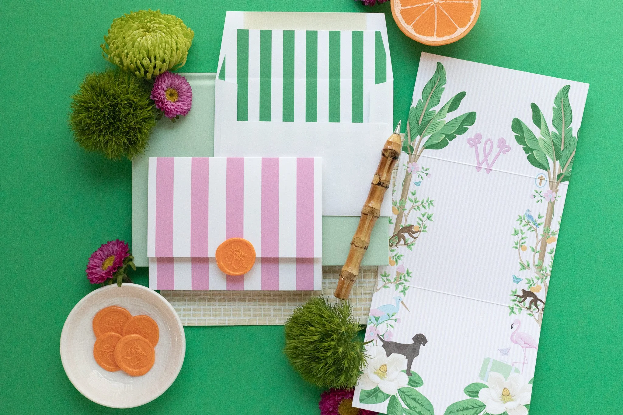 Stationery and decorative items on a green background, including striped envelopes, a botanical illustrated paper, a bamboo pen, green flowers, pink and purple flowers, a half orange, and a white dish with orange wax seals.
