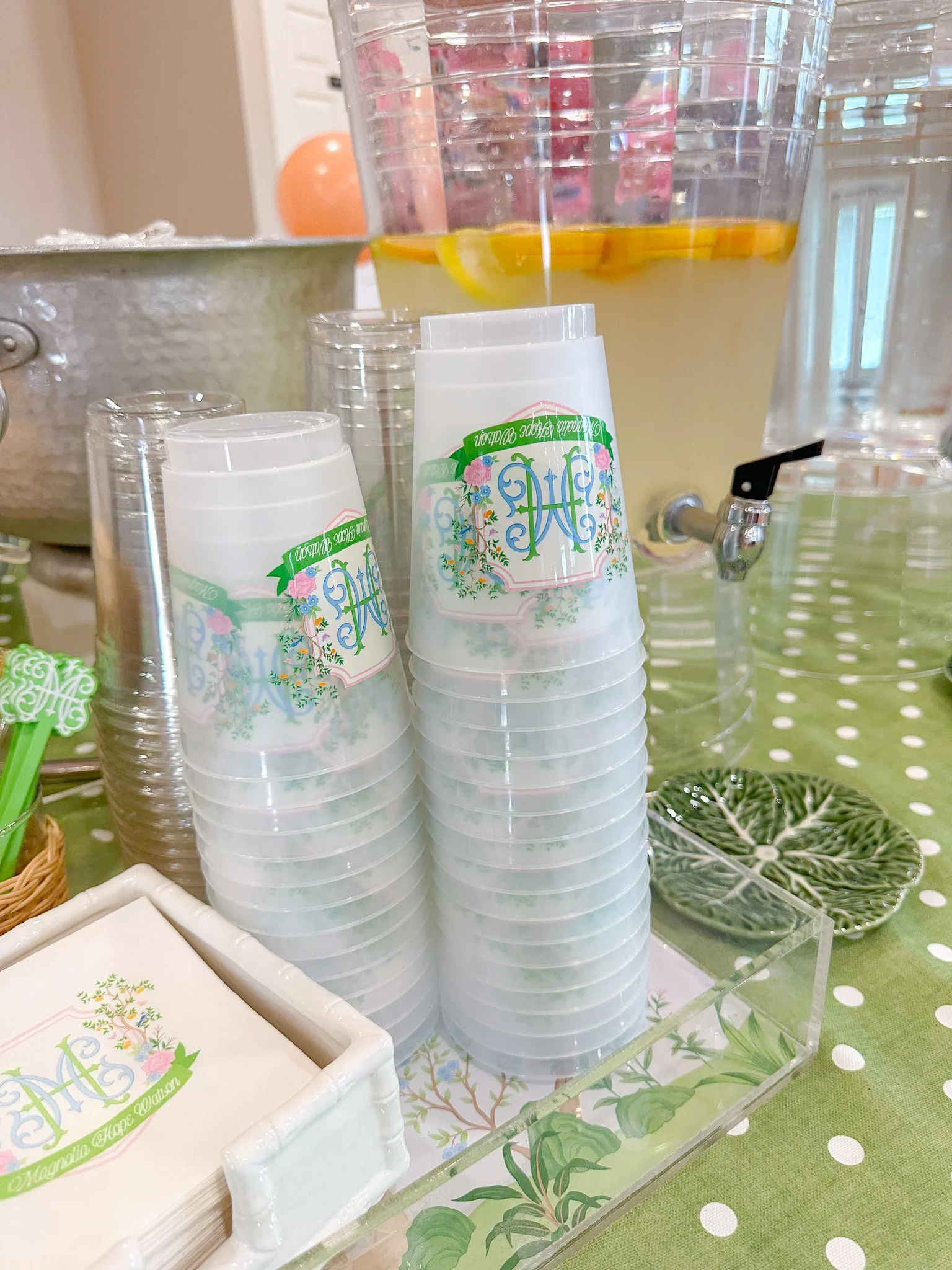 Stacked clear plastic cups with floral and monogram design, lemonade with lemon slices in a clear drink dispenser, and a leaf-shaped plate on a polka dot green tablecloth.