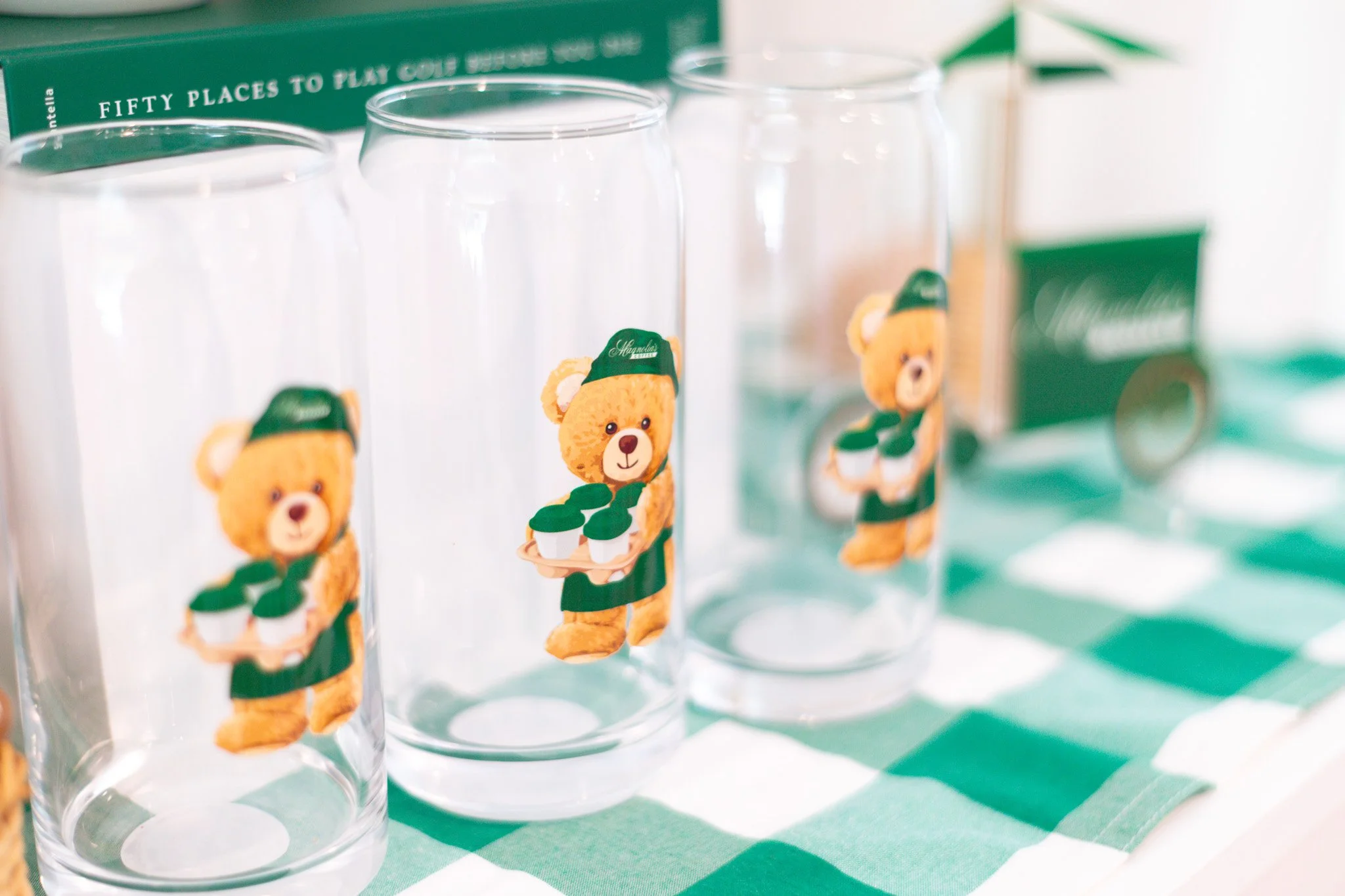 Clear glasses with a teddy bear carrying a tray of plants sticker, arranged on a green and white checkered tablecloth, with a green and white cone-shaped umbrella in the background.