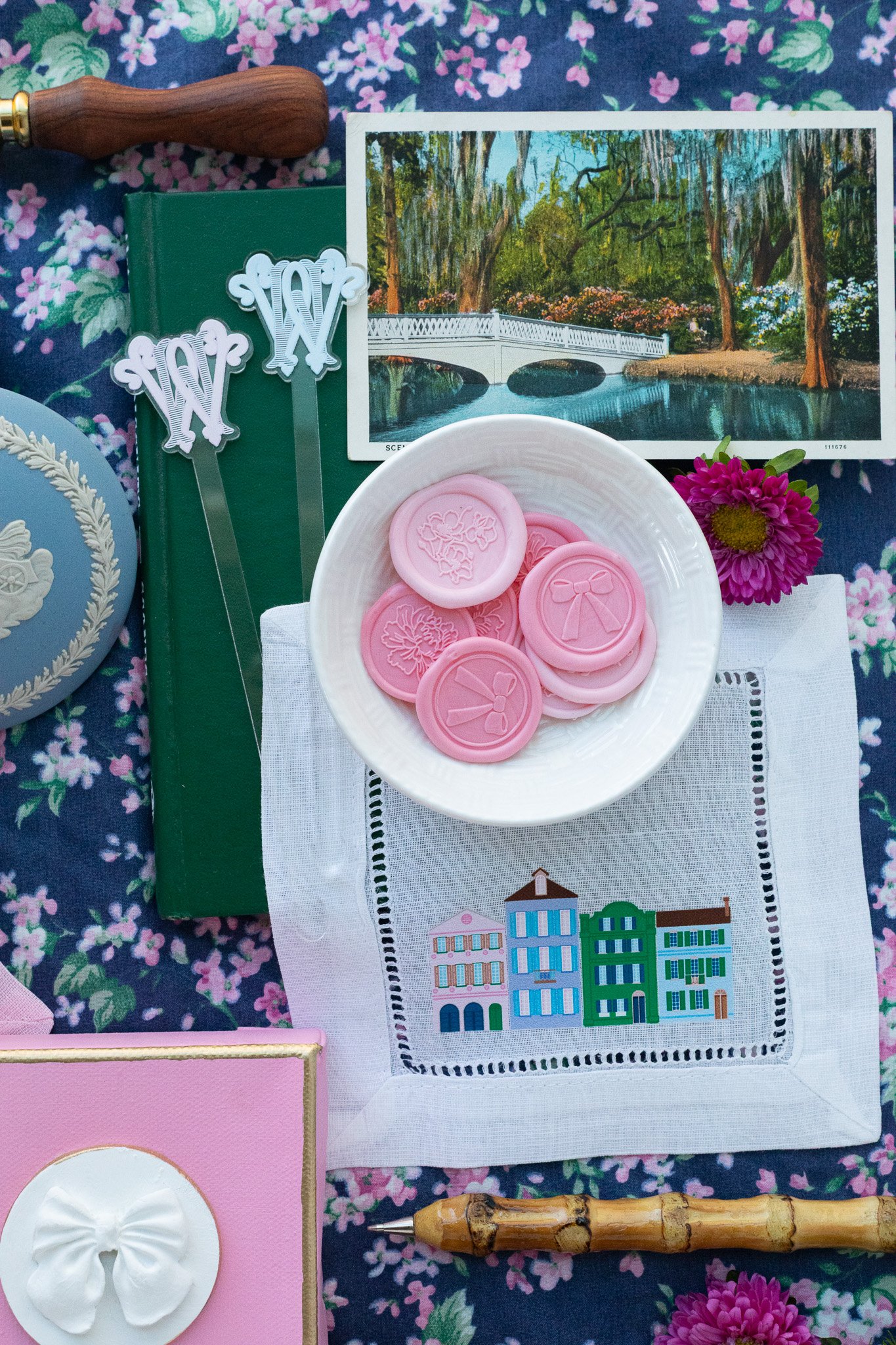 Pink wax seals with holiday and bow designs in a white bowl on a cloth with colorful houses embroidery, surrounded by a pink box, a flower, a postcard of a park, two pins, a wax seal stamp with a wooden handle, and a floral tablecloth.