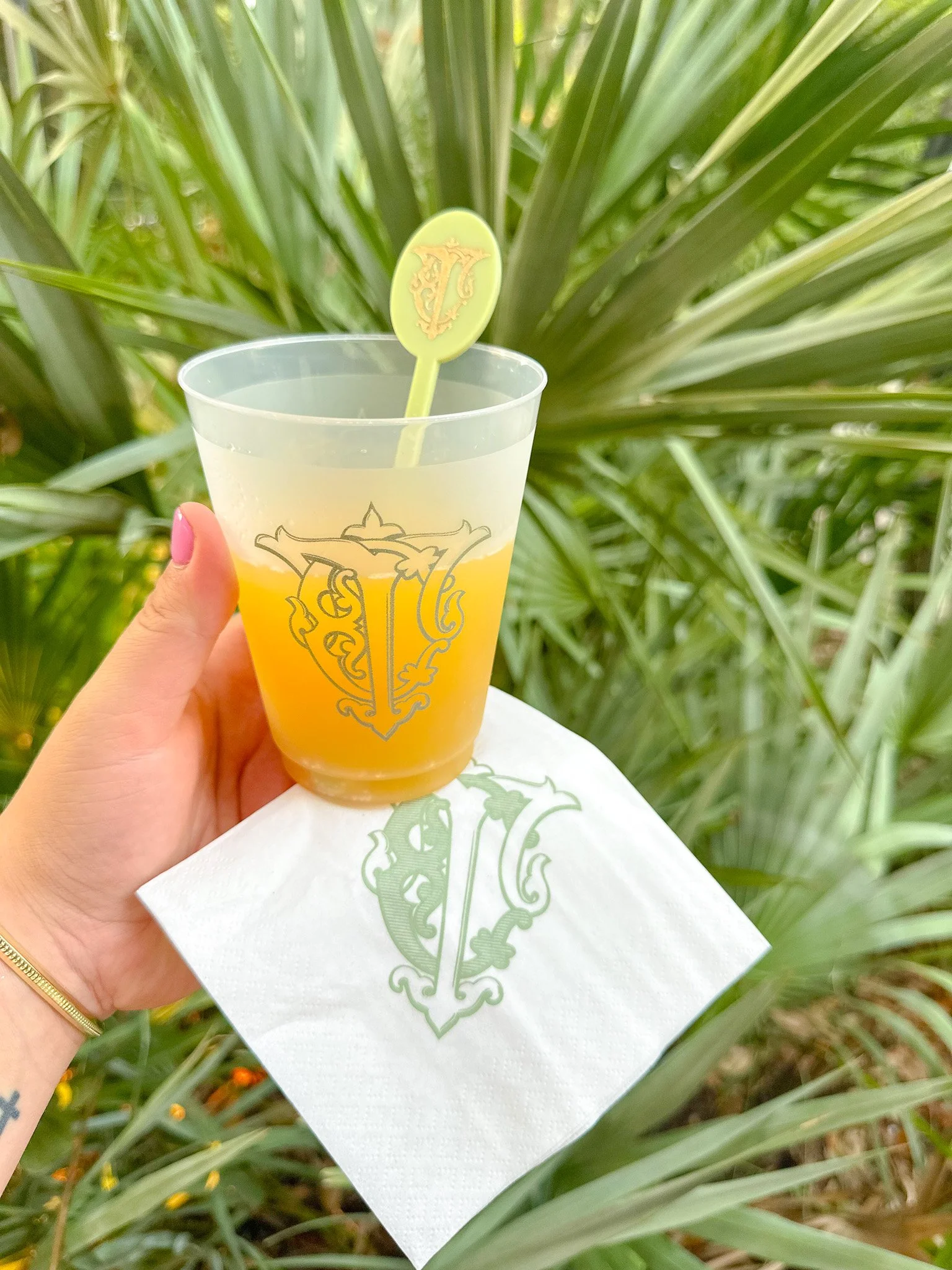A hand holding a clear plastic cup filled with orange juice and a green stir stick, with a white napkin underneath it, outdoors among tall green plants.