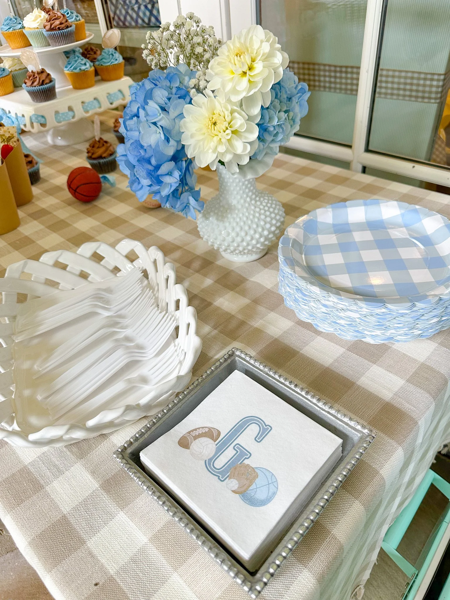 Table decorated with a blue and white checkered theme, featuring a bouquet of white and blue flowers in a textured white vase, stacked blue and white checkered plates, with white plastic utensils and a silver tray holding paper napkins with a ball an