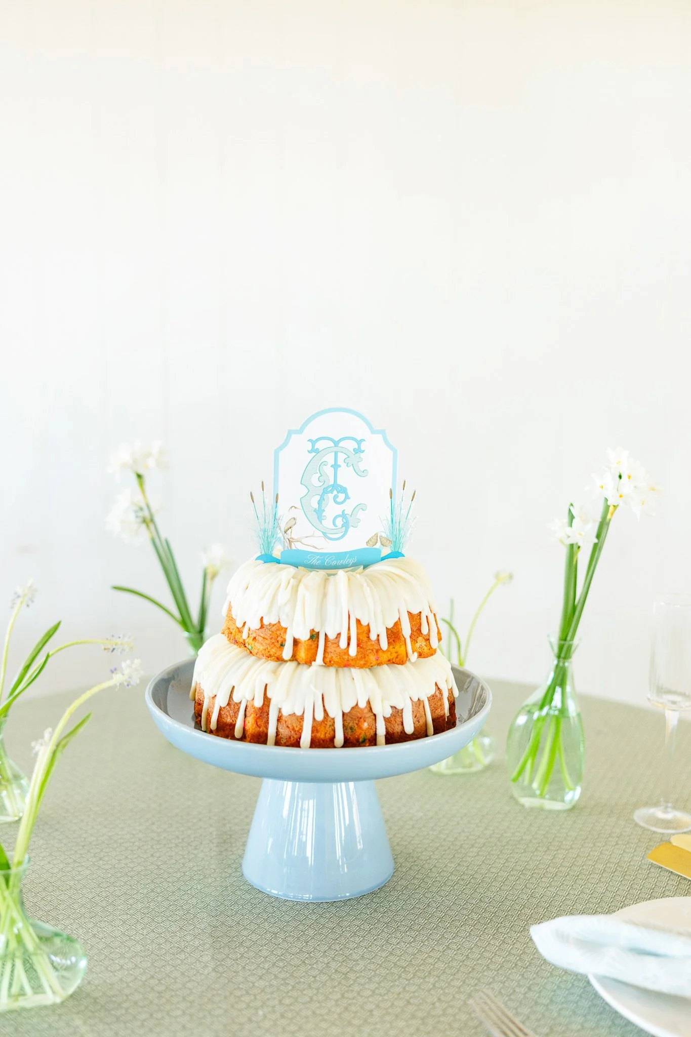 Two-tiered cake with white frosting, white chocolate drizzle, and decorative topper, on a light blue cake stand, surrounded by small vases with white flowers on a table.