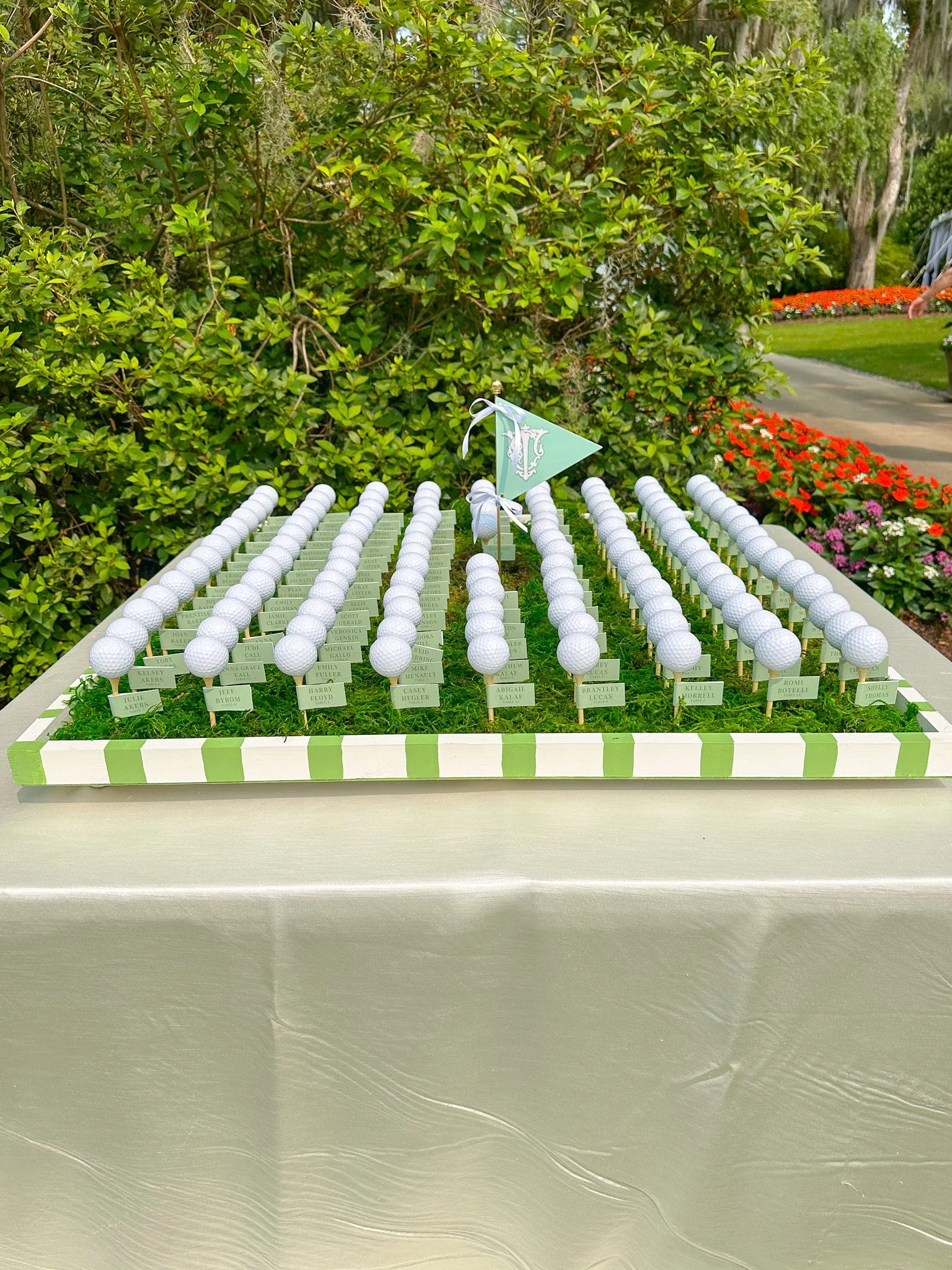 A display of golf balls with small nameplates beneath each, set on a table with a green and white striped border, in an outdoor garden setting. There is a small triangular flag with a letter 'T' on it at the center of the display.