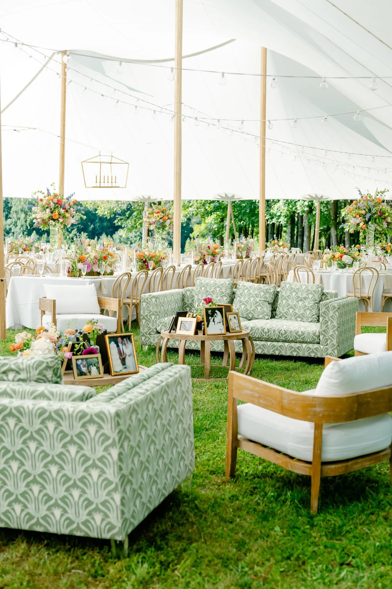 Decorated outdoor wedding reception tent with floral arrangements, wooden chairs, and framed photographs on a green lawn.