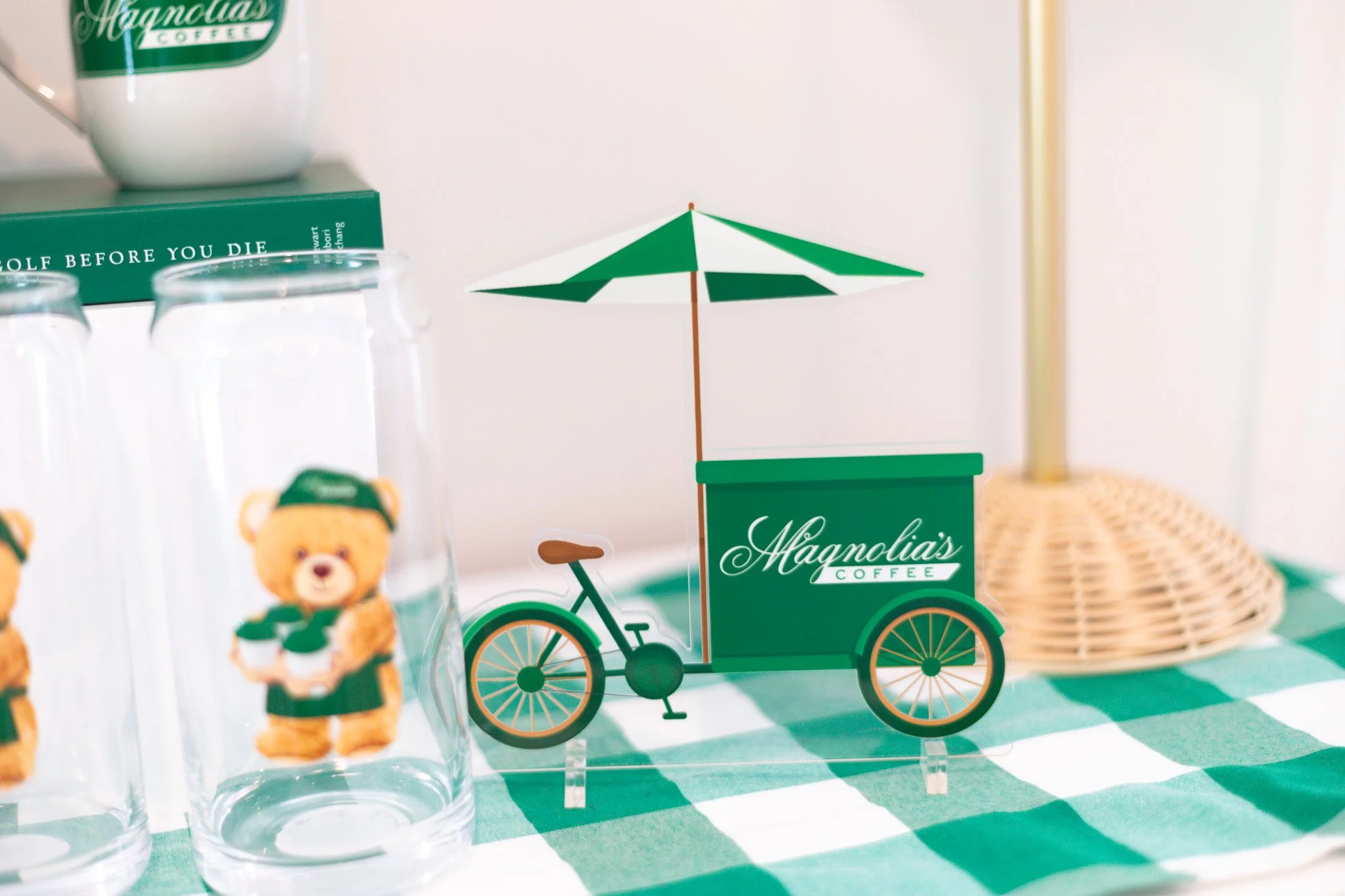 Decorative display with a Magnolia's Coffee sign, a miniature bicycle with a coffee cart, cups of Teddy Bear Pee-wee glasses, a green checkered tablecloth, a green and white umbrella, and a wicker basket in the background.