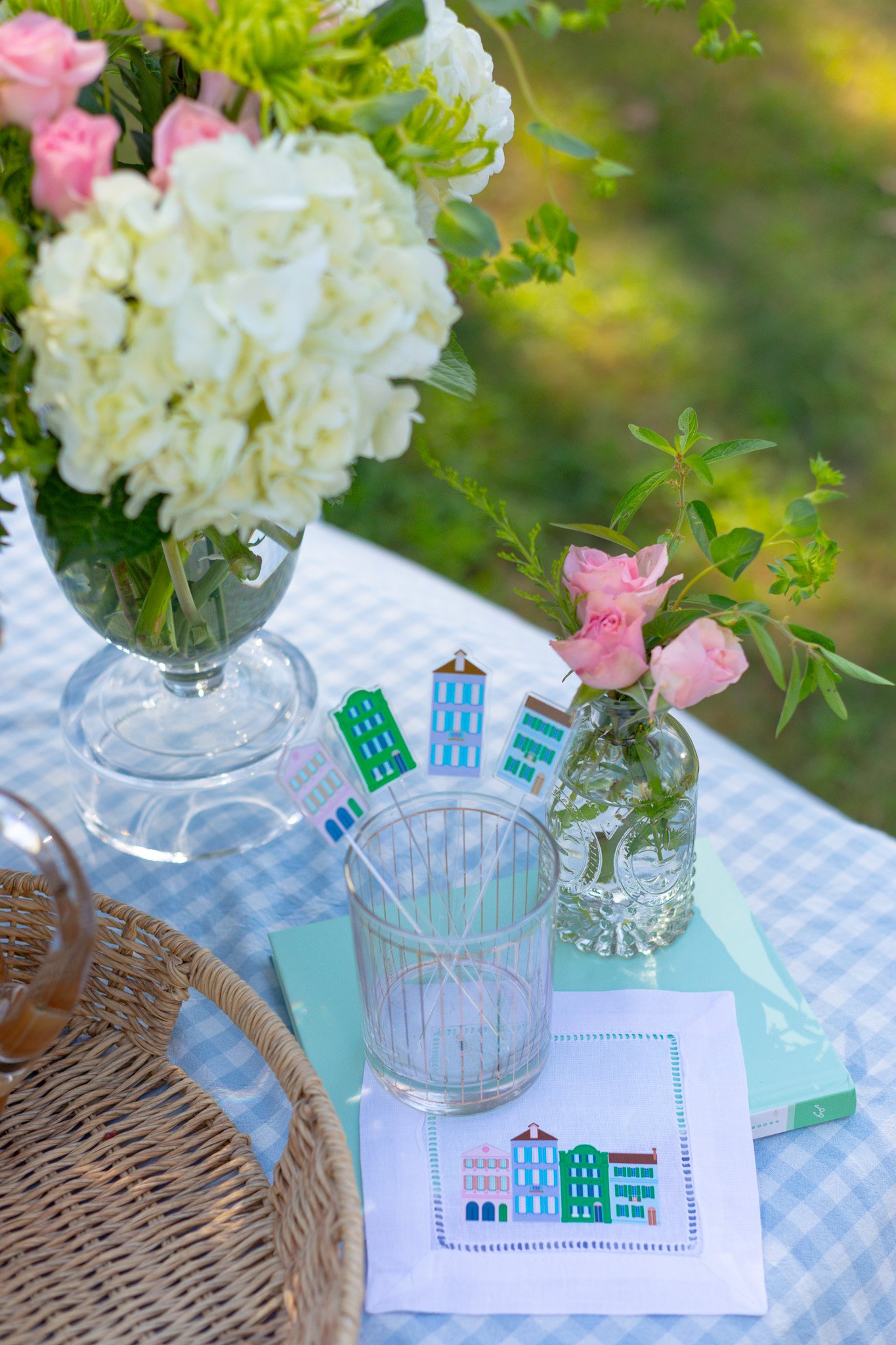 A table with a blue-checkered tablecloth, decorated with two vases of pink and white flowers, a glass with colorful city skyline-themed cupcake toppers, a book, a cloth napkin with city buildings design, and a woven basket, set outdoors.