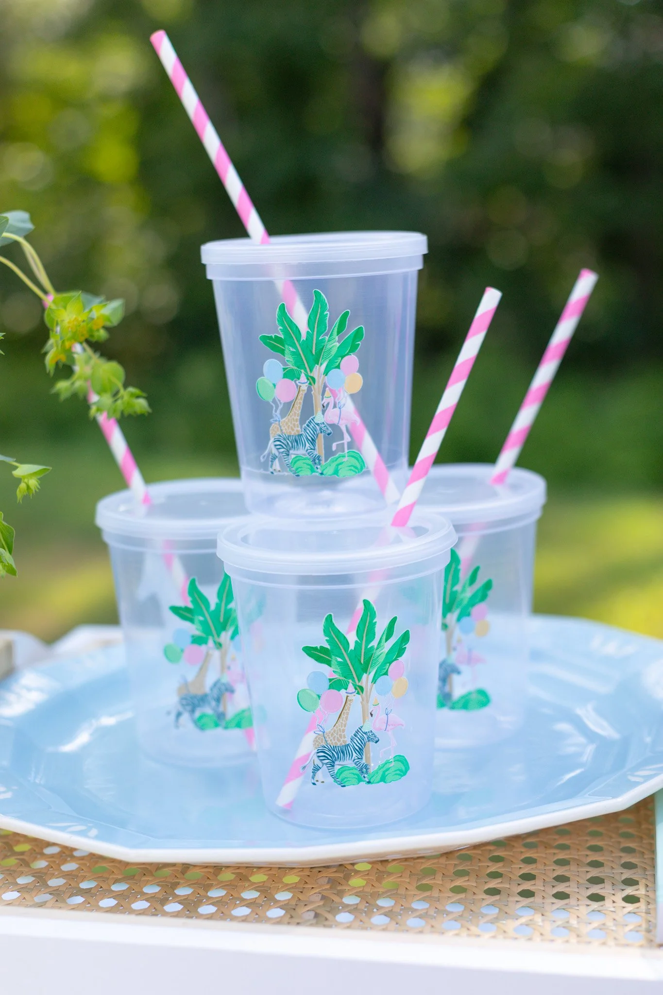 Four clear plastic cups with pink and white striped straws and a jungle-themed design featuring trees, zebras, giraffes, and balloons, sitting on a white tray outdoors.