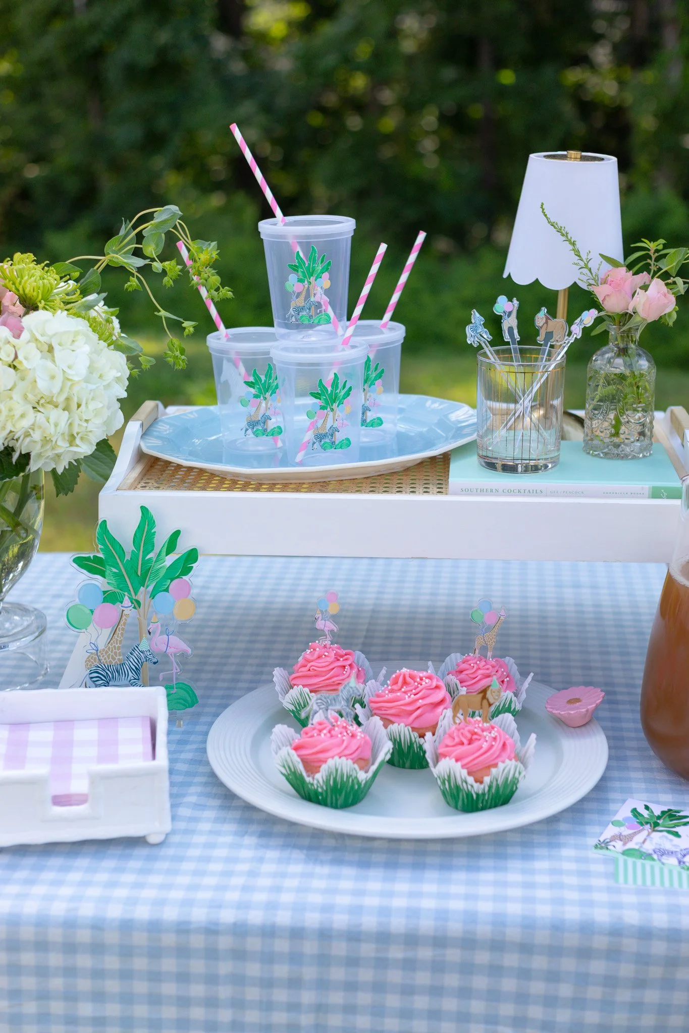 Table set with pink cupcakes, clear cups with animal decorations, straws, flowers, and a lamp at an outdoor party.