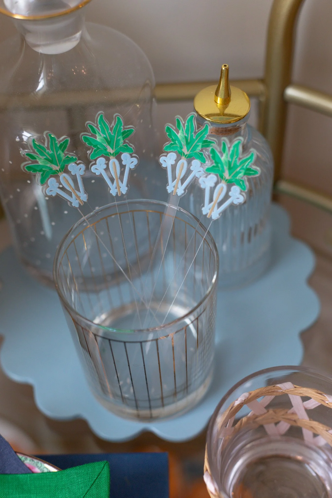 Clear glass water bottle with green and white plant-like stickers, a small clear glass with gold rimmed lid, and a glass with a gold rim, all placed on a light blue scalloped tray.