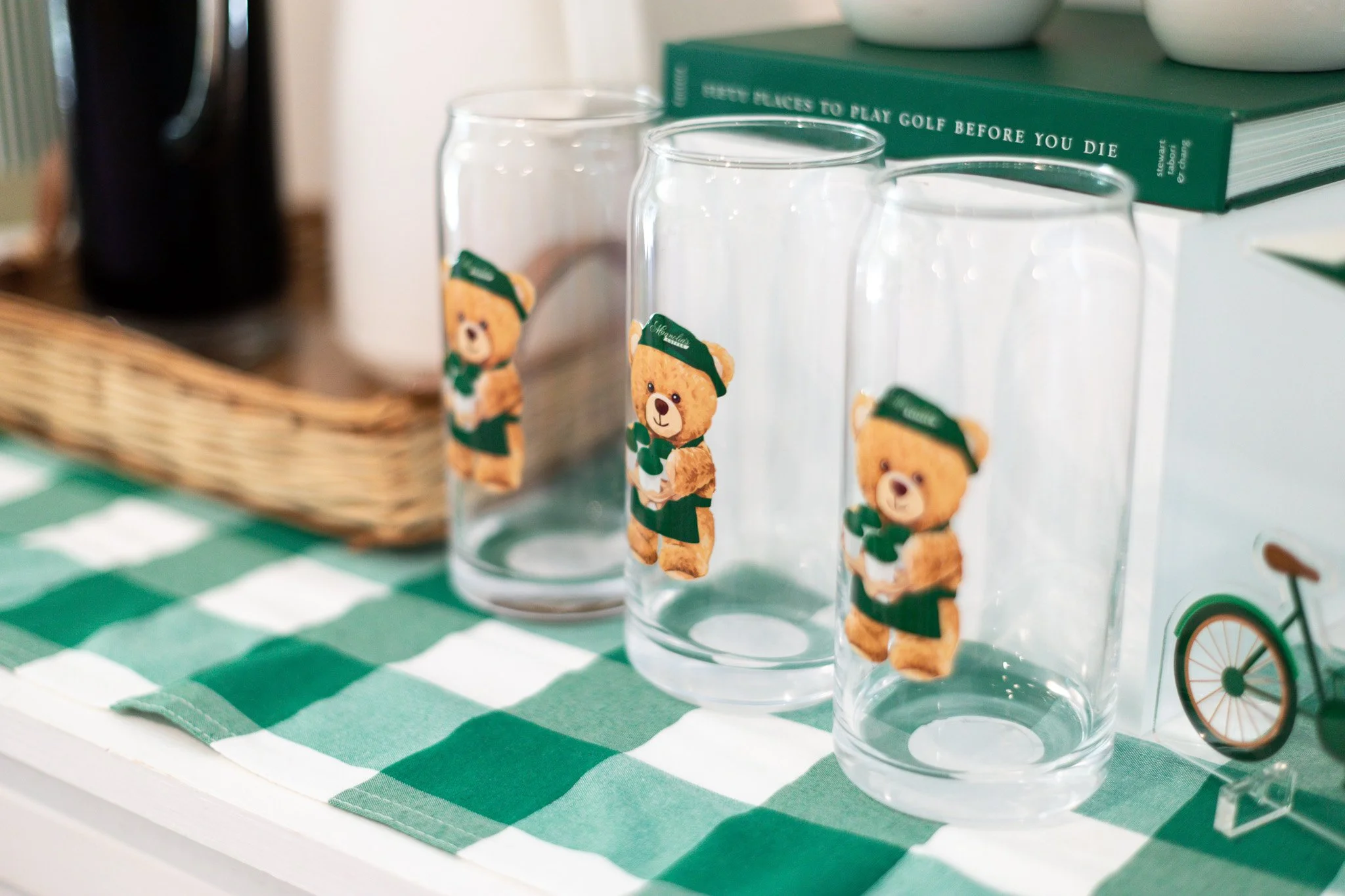 Three clear glass tumblers with bear stickers, placed on a green and white checkered cloth, with a basket and stacked books in the background.