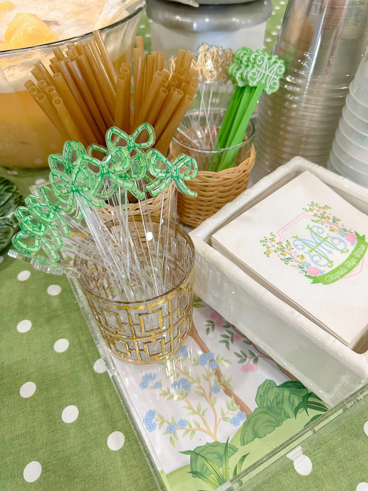 Party table setup with green and floral themed napkins, clear and decorated cups holding green bow and monogram stir sticks, a basket with green floral napkins, clear plastic cups stacked, and containers with straws and utensils on a green polka-dott