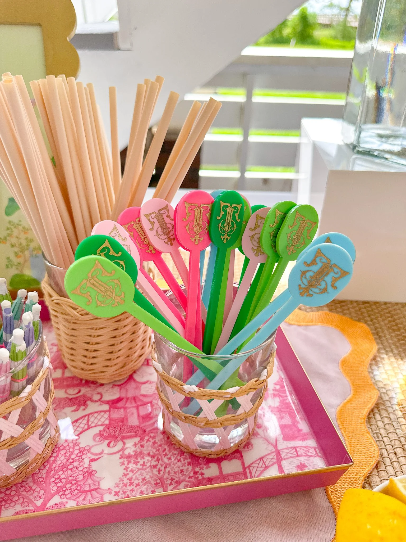 Colorful plastic spoons and straw stirrers in glass and wicker containers on a pink tray with a Fairy Tale or amusement park print, set on a table near a window.