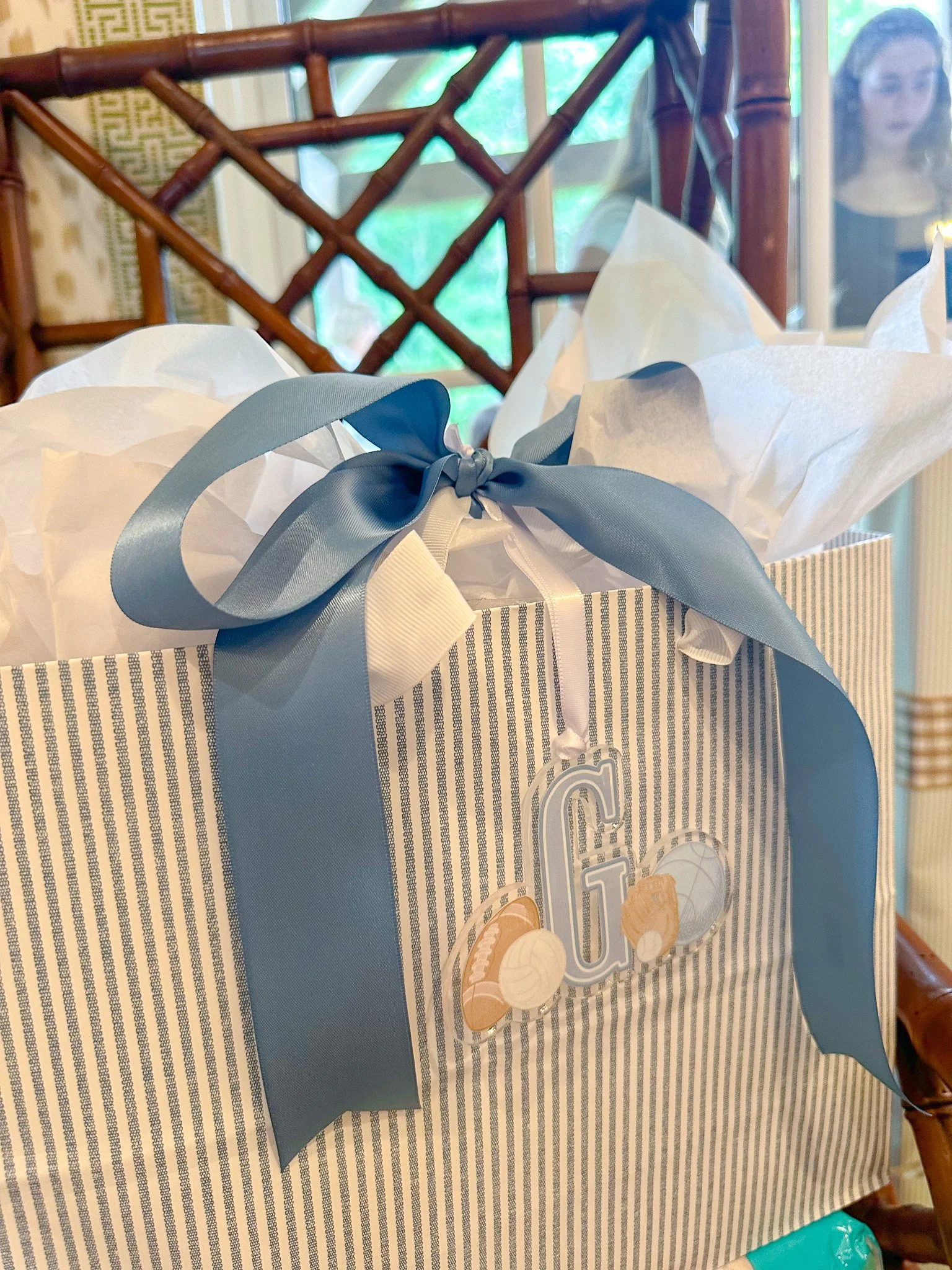 Close-up of a striped gift bag decorated with a wide blue ribbon bow and white tissue paper, with an acrylic monogrammed letter 'G' and football-themed embellishments attached to the front.