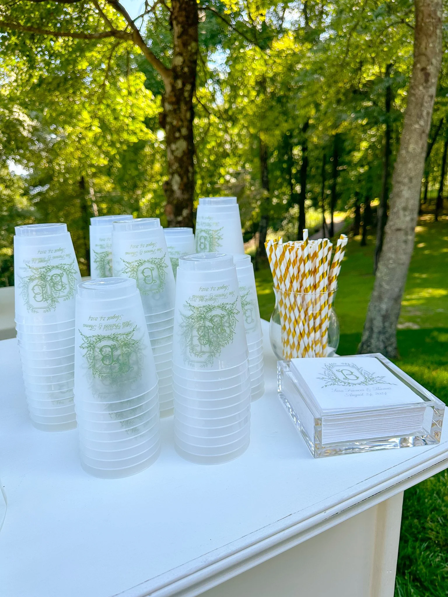 A white table outdoors with stacks of clear plastic cups, some with a green decorative design and text, a container holding yellow and white striped paper straws, and a clear acrylic tray with napkins featuring a decorative monogram and text. The bac