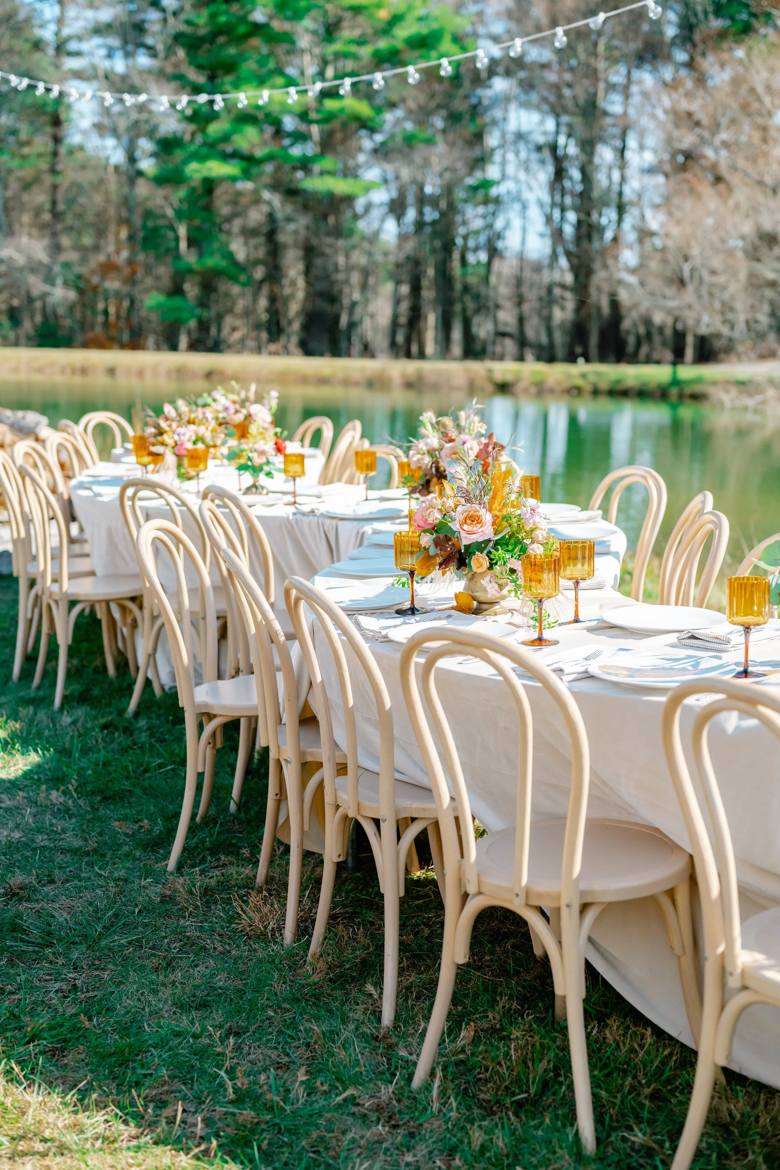 Outdoor dining table set with floral centerpieces and amber glasses near a pond, with string lights overhead and trees in the background.
