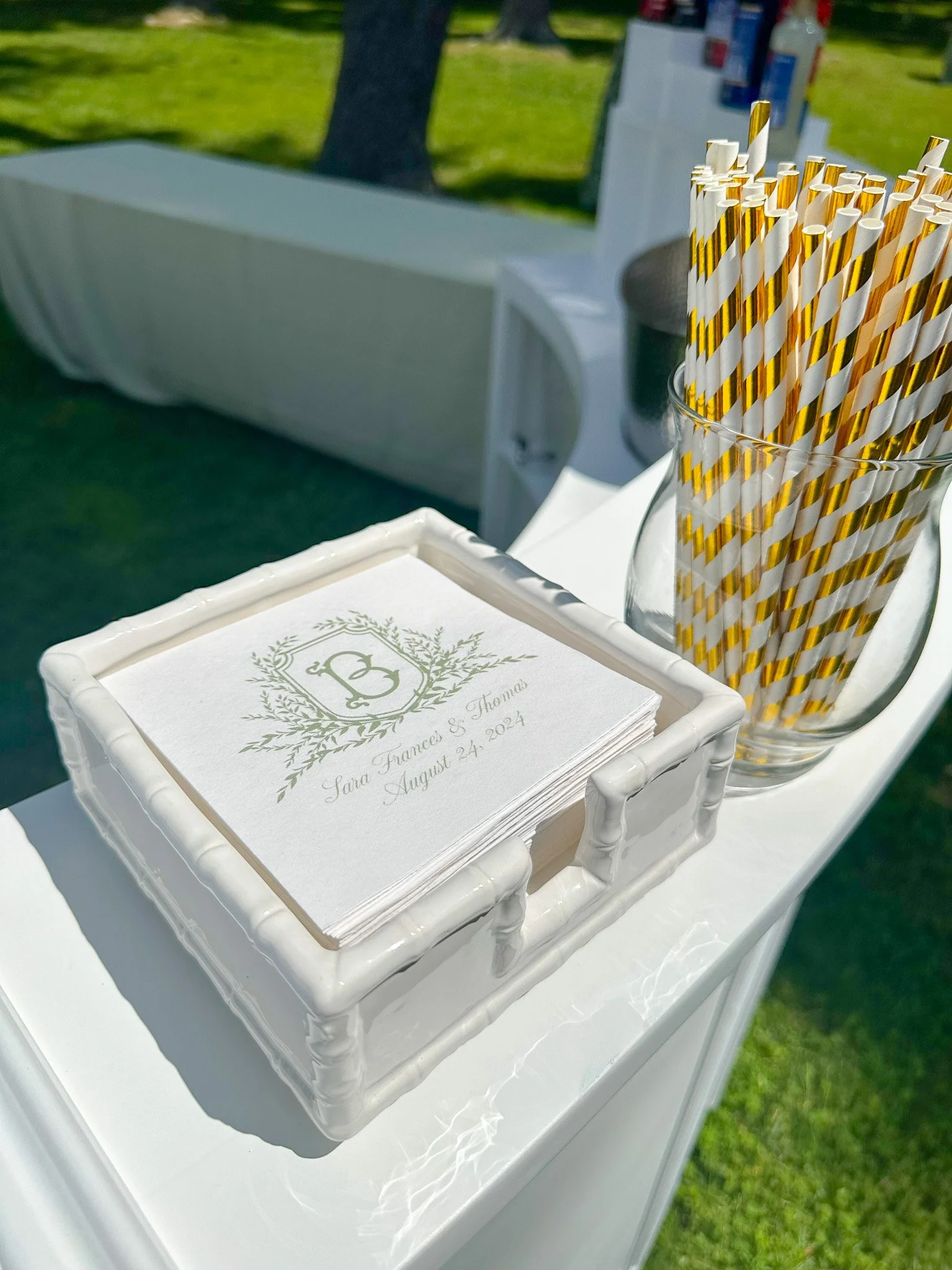 Wedding program with the names Sara Frances and Thomas, dated August 24, 2024, on a white tray, next to a glass jar filled with gold and white striped paper straws, outdoors on a sunny day.