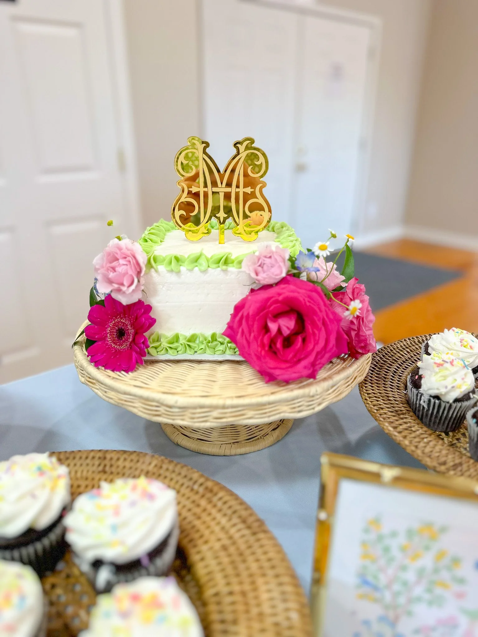 A white birthday cake with green icing border, decorated with pink and purple flowers, topped with a gold and pink monogram cake topper, placed on a wicker cake stand. Surrounding the cake are cupcakes with white frosting and colorful sprinkles.