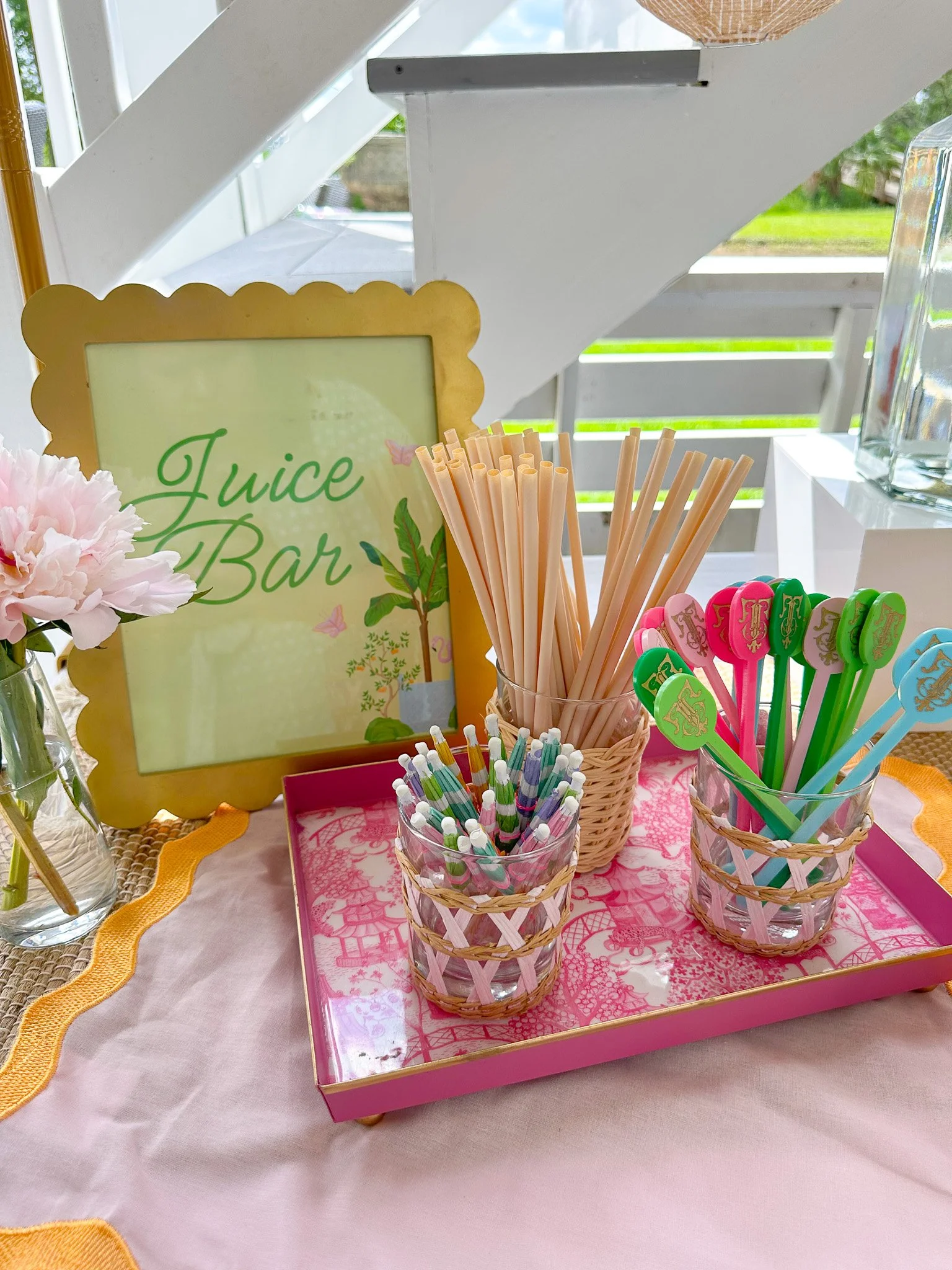 Juice bar setup with pink tray containing colorful straws, pink, green, and blue spoons, a sign saying 'Juice Bar', and a vase with pink flowers.