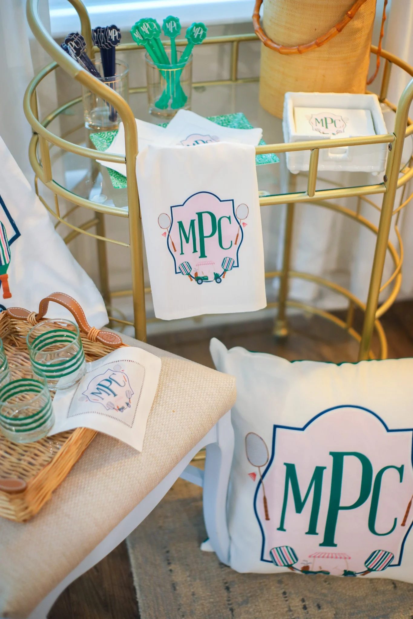 A display of T-shirts, a pillow, napkins, and cups with a logo that includes the initials 'MPC' and illustrations of tennis rackets and paddles, arranged on a table and a gold cart, likely for a tennis-themed event or party.