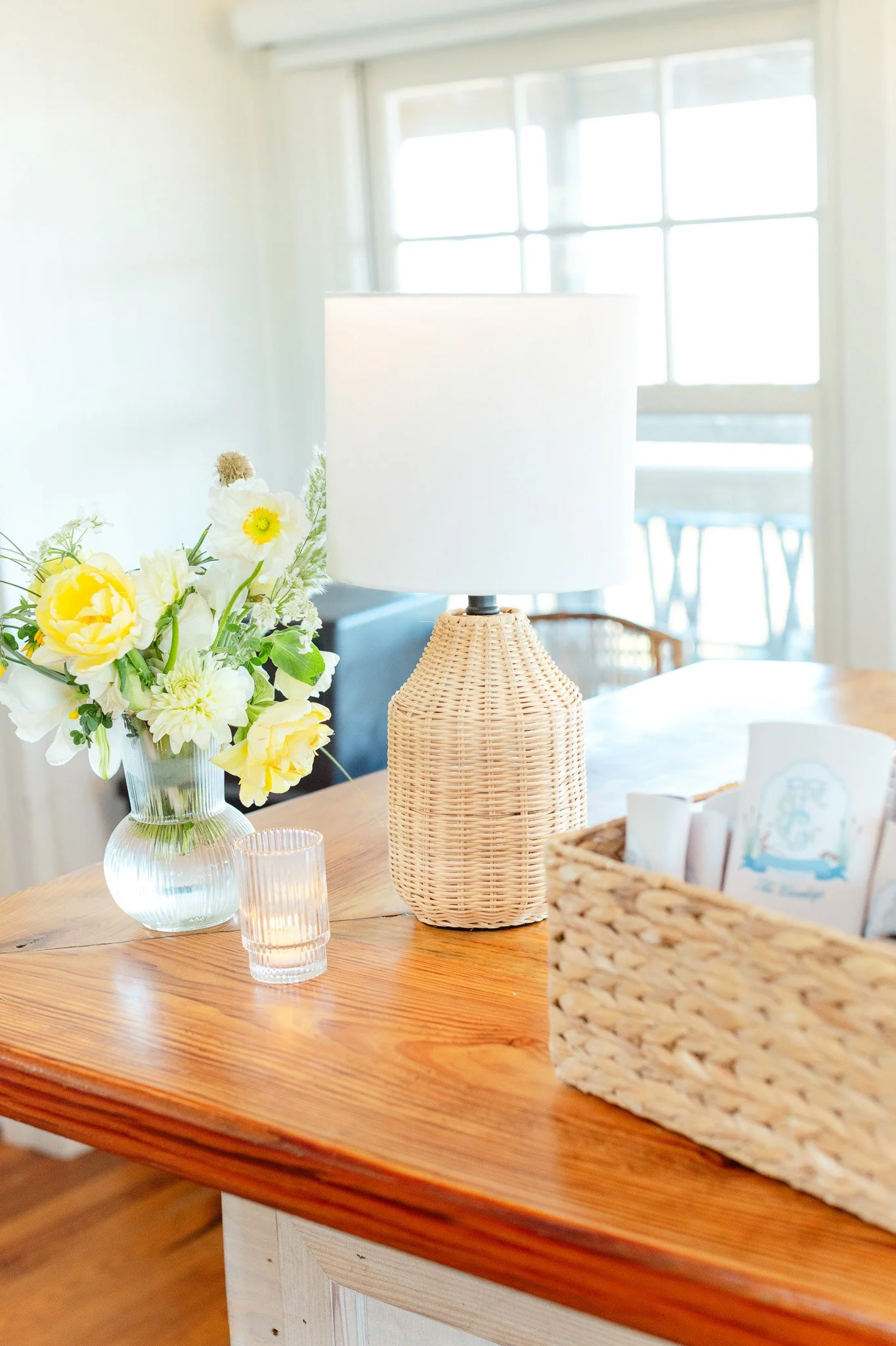 A wooden table with a white lampshade, a glass vase of yellow and white flowers, a small glass candle holder, and a woven basket with papers inside. A large window in the background lets in natural light.