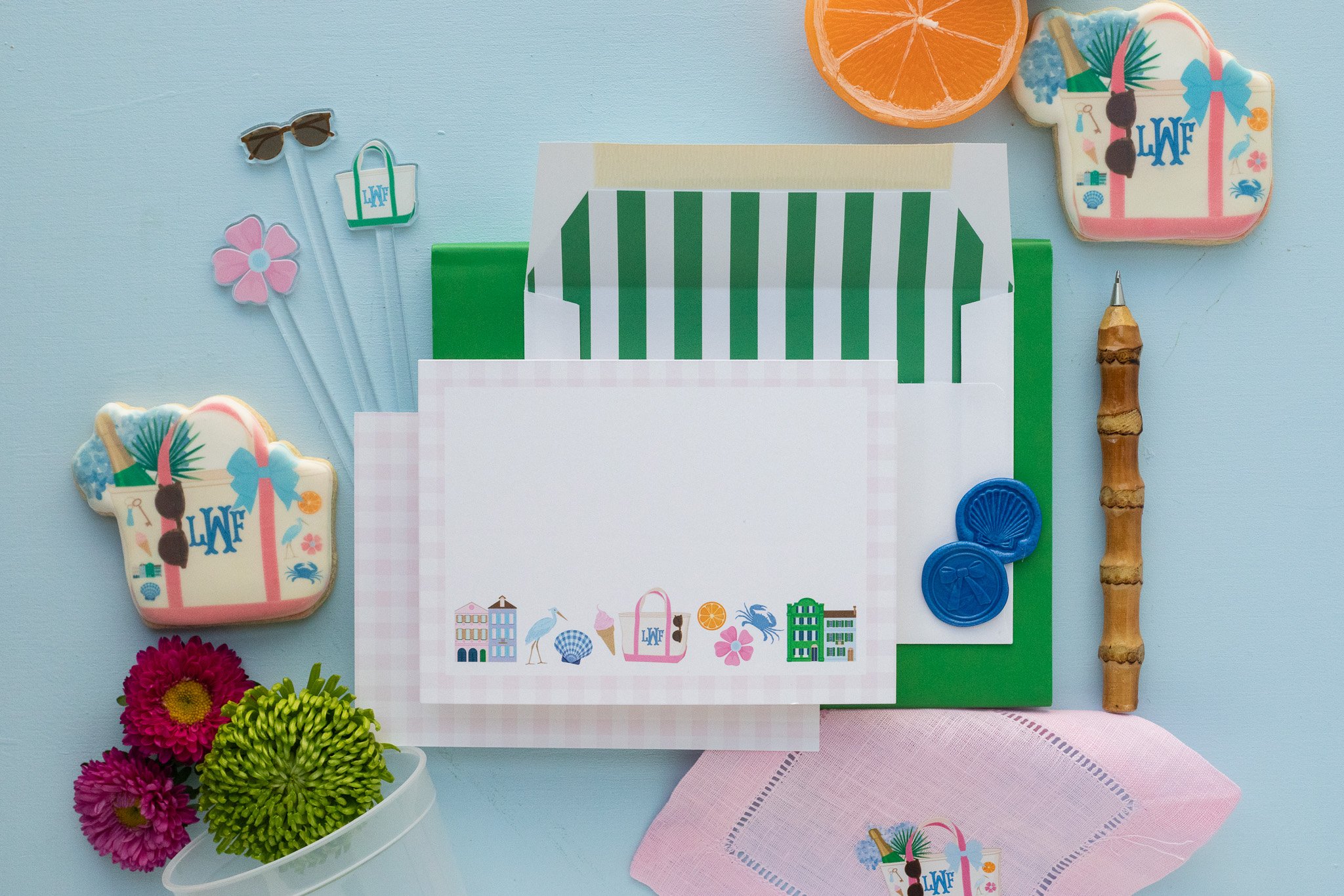 Beach-themed stationery and accessories on a light blue surface, including a floral napkin, pink and green flowers, ice cream and beach bag stickers, a bamboo stick, pink sunglasses, pink and green folders, and a sliced orange.