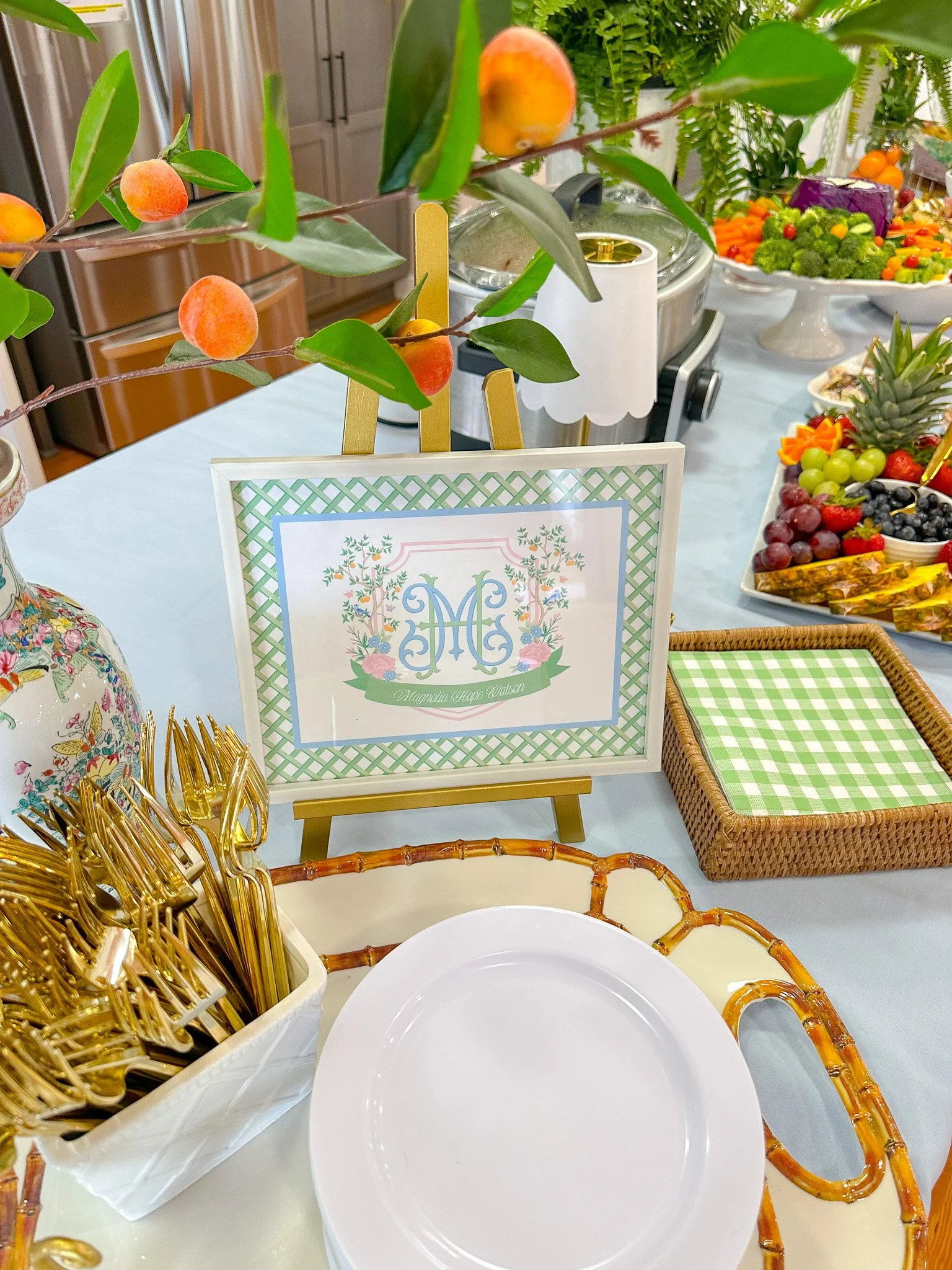 Table decorated for a party or event with a sign featuring monogrammed initials 'MC' and floral design, surrounded by gold utensils, a bamboo tray, and a fruit platter with grapes, berries, and tropical fruit in the background.