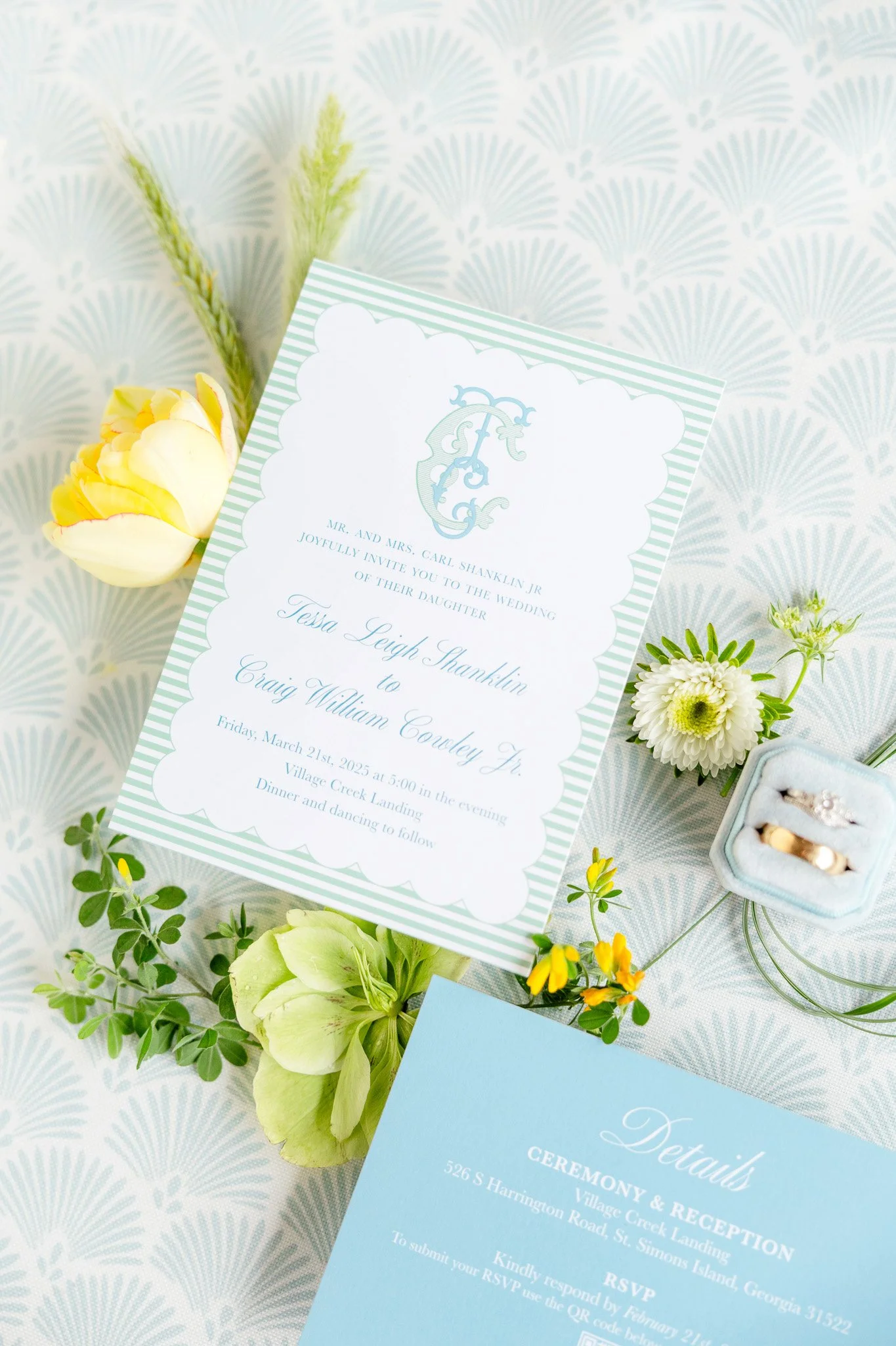 A wedding invitation card, a blue details card, a ring box with rings, and flowers on a patterned tablecloth.