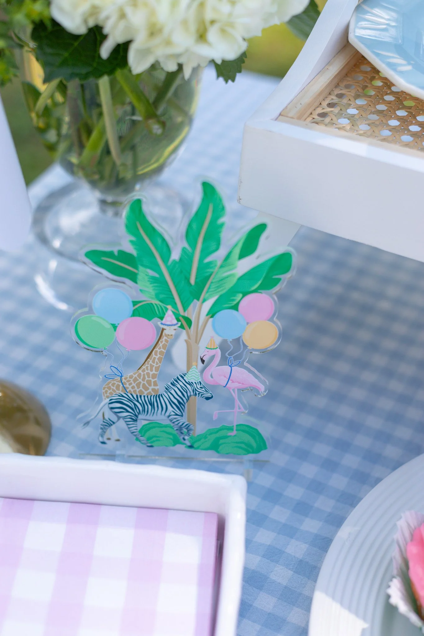 Decorative paper cutout featuring a zebra, giraffe, and flamingo with balloons and a palm tree, placed on a table at a celebration.