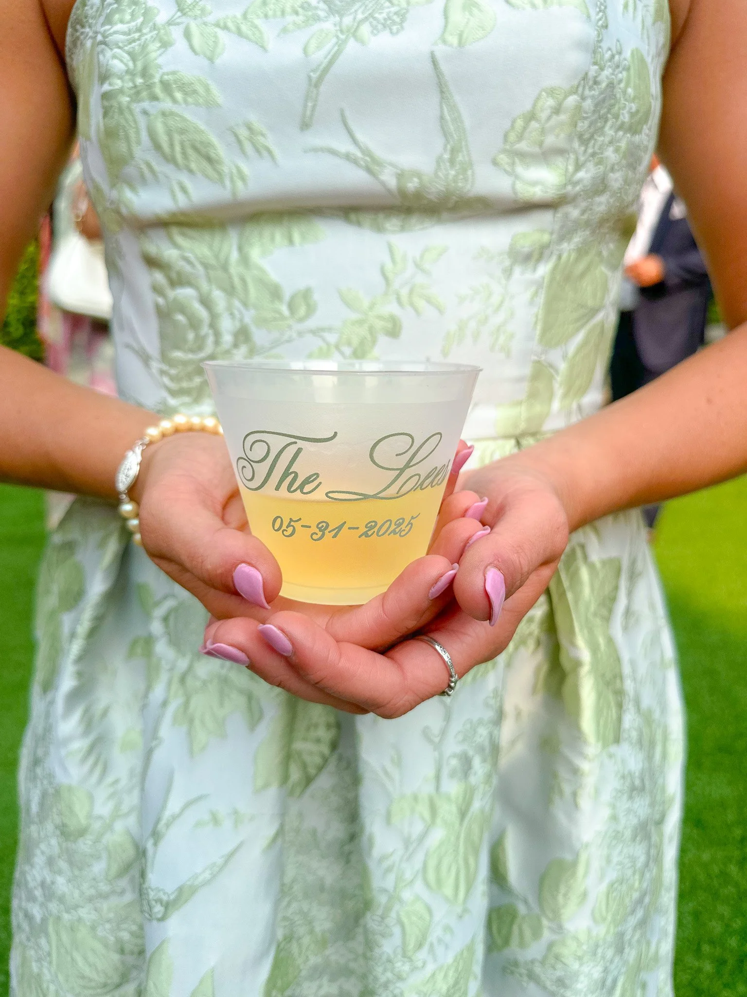 Person in a white and green floral dress holding a cup with the text 'The Lord' and a date '05-31-2023' written on it, at outdoor event.