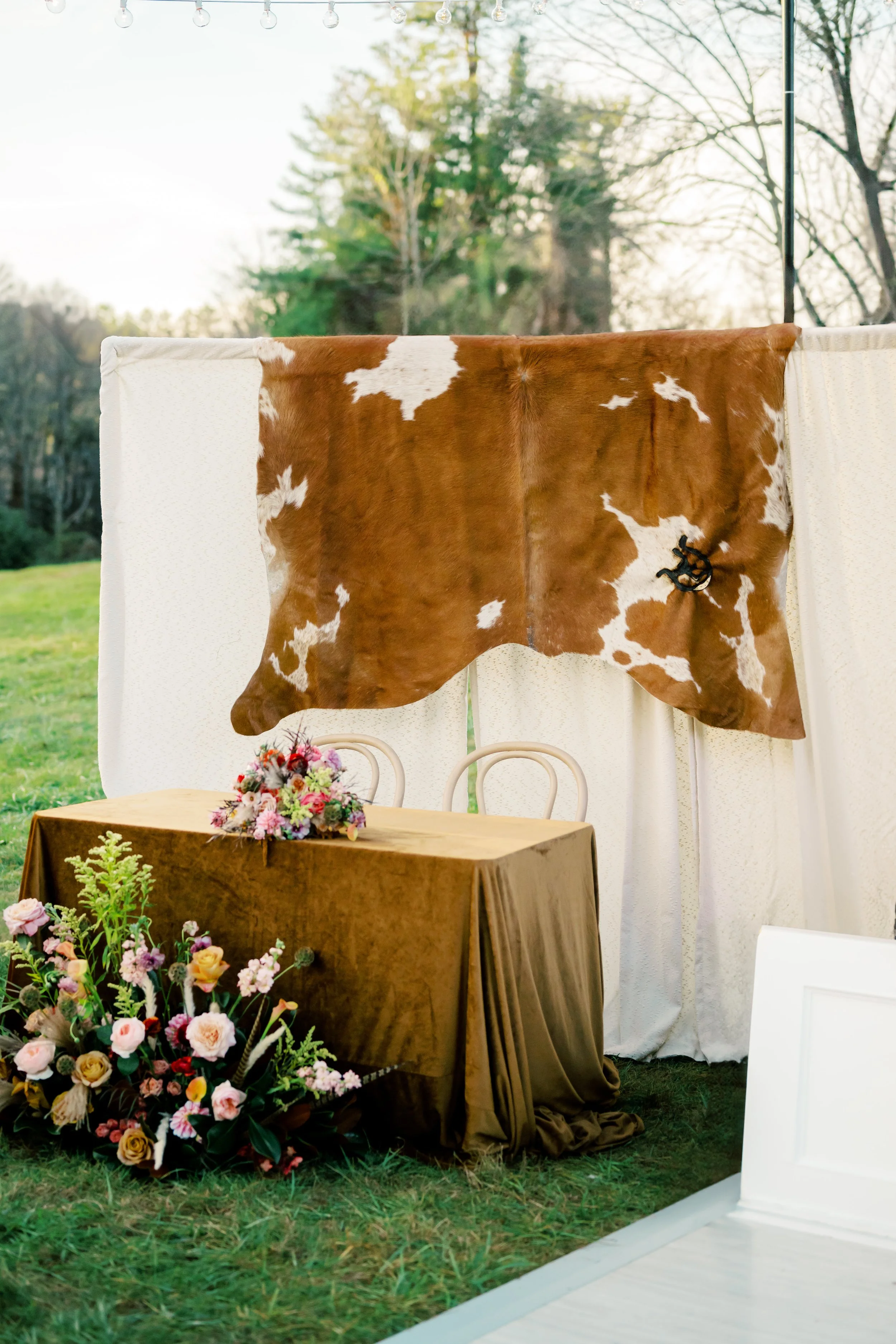 Table decorated with flowers in front of a rustic cowhide curtain outdoors on grass.