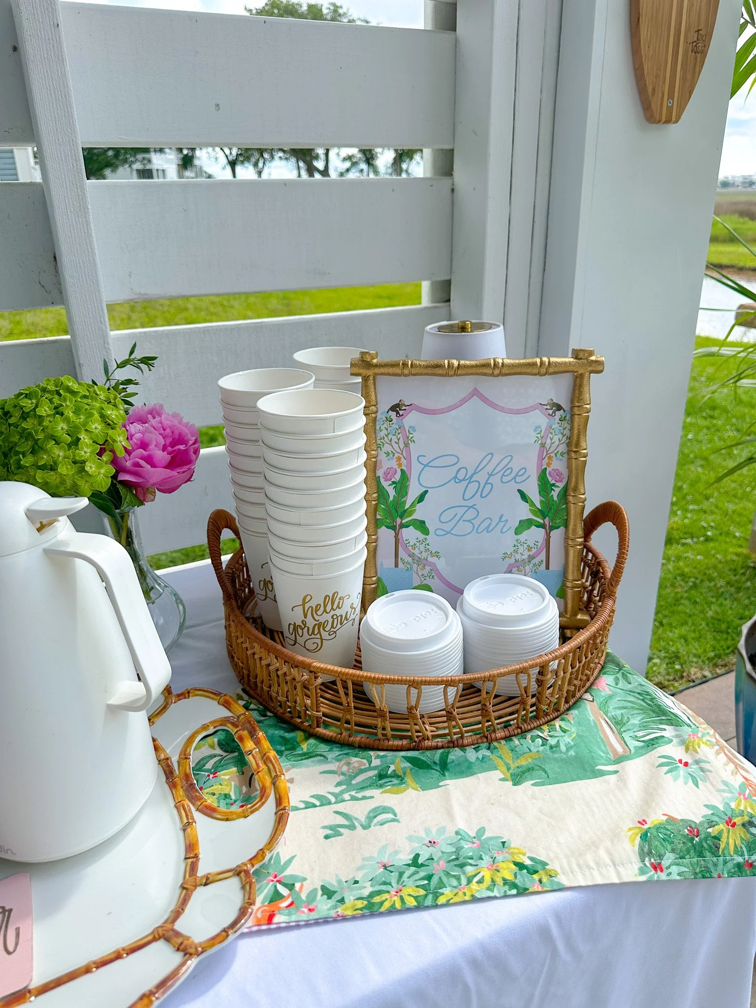 A coastal-themed coffee bar setup outdoors with a wicker basket holding white disposable cups, in front of a decorative sign that reads 'Coffee Bar,' and a white electric kettle on a floral tablecloth with a pink peony and green hydrangea flowers nea