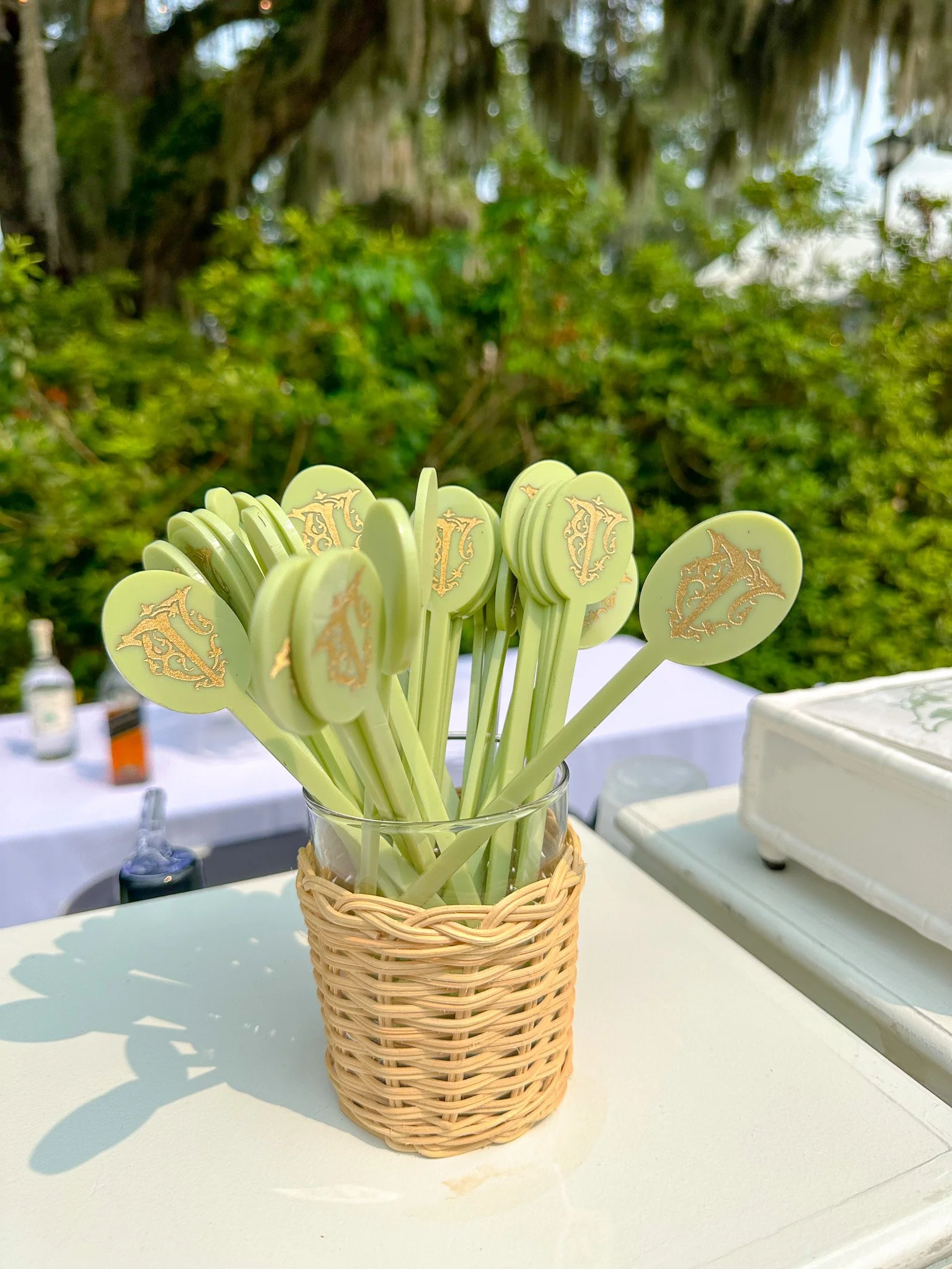Green plastic spoons with gold writing in a glass holder wrapped with a wicker cover, set on a white outdoor table with a shaded garden background.