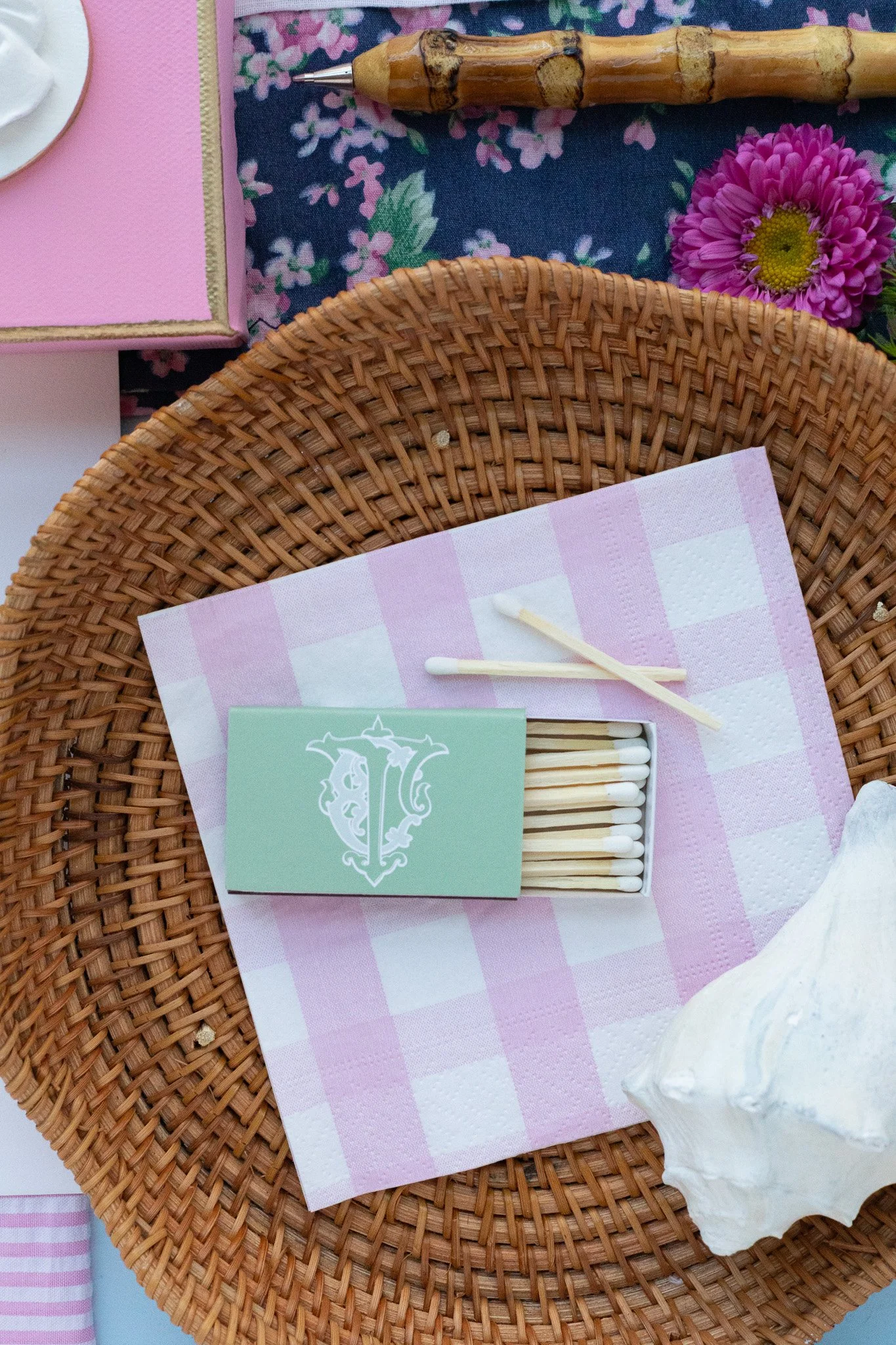 A wicker basket lined with a pink and white checkered napkin holding a small green box of matches. Two matchsticks rest on the napkin. Nearby, there is a large seashell, a pink flower, and part of a pink box with a gold zipper. At the top, a bamboo p
