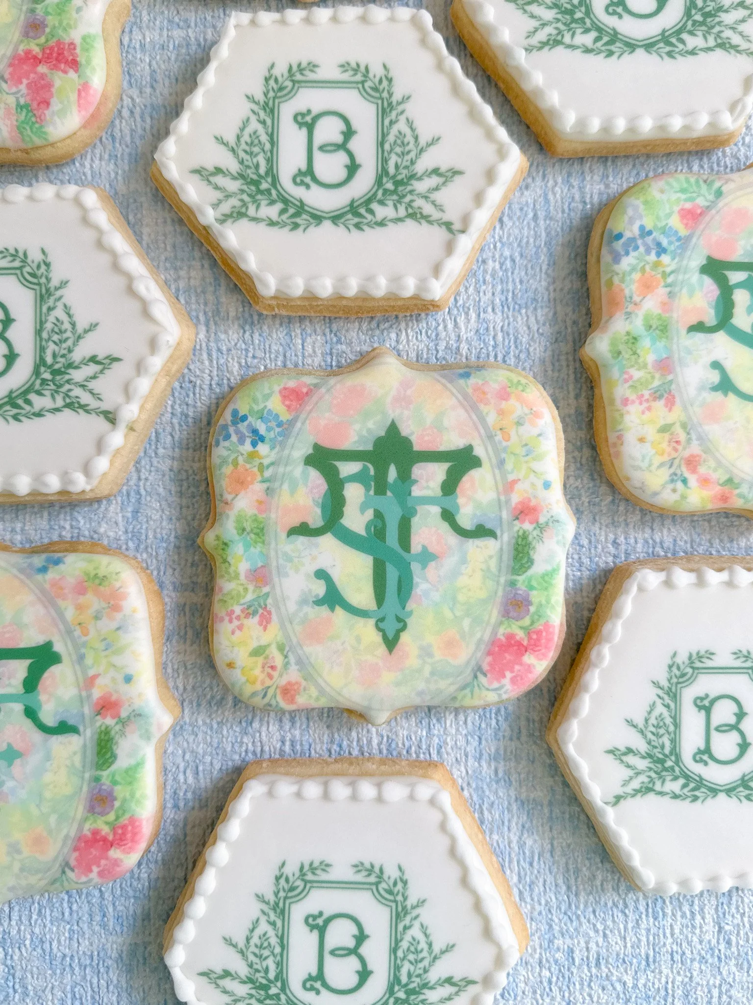 Decorated cookies with white icing and monogram designs, some with green script letters and floral patterns, arranged on a blue textured surface.