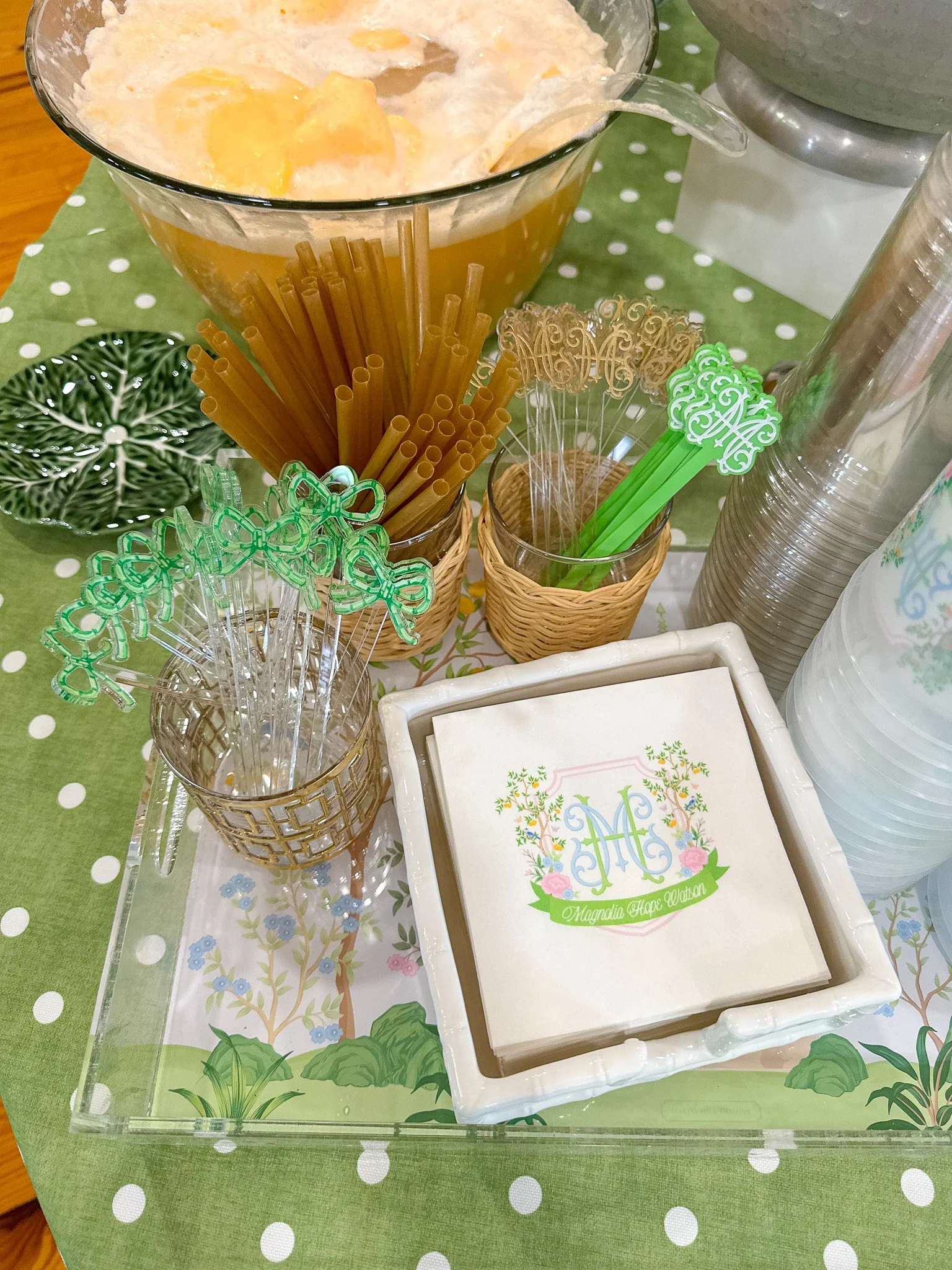 A table setup with a large glass bowl of vegetable soup, pasta, and a variety of cocktail stirrers, napkins with a decorative logo, and disposable cups, on a green polka dot tablecloth.