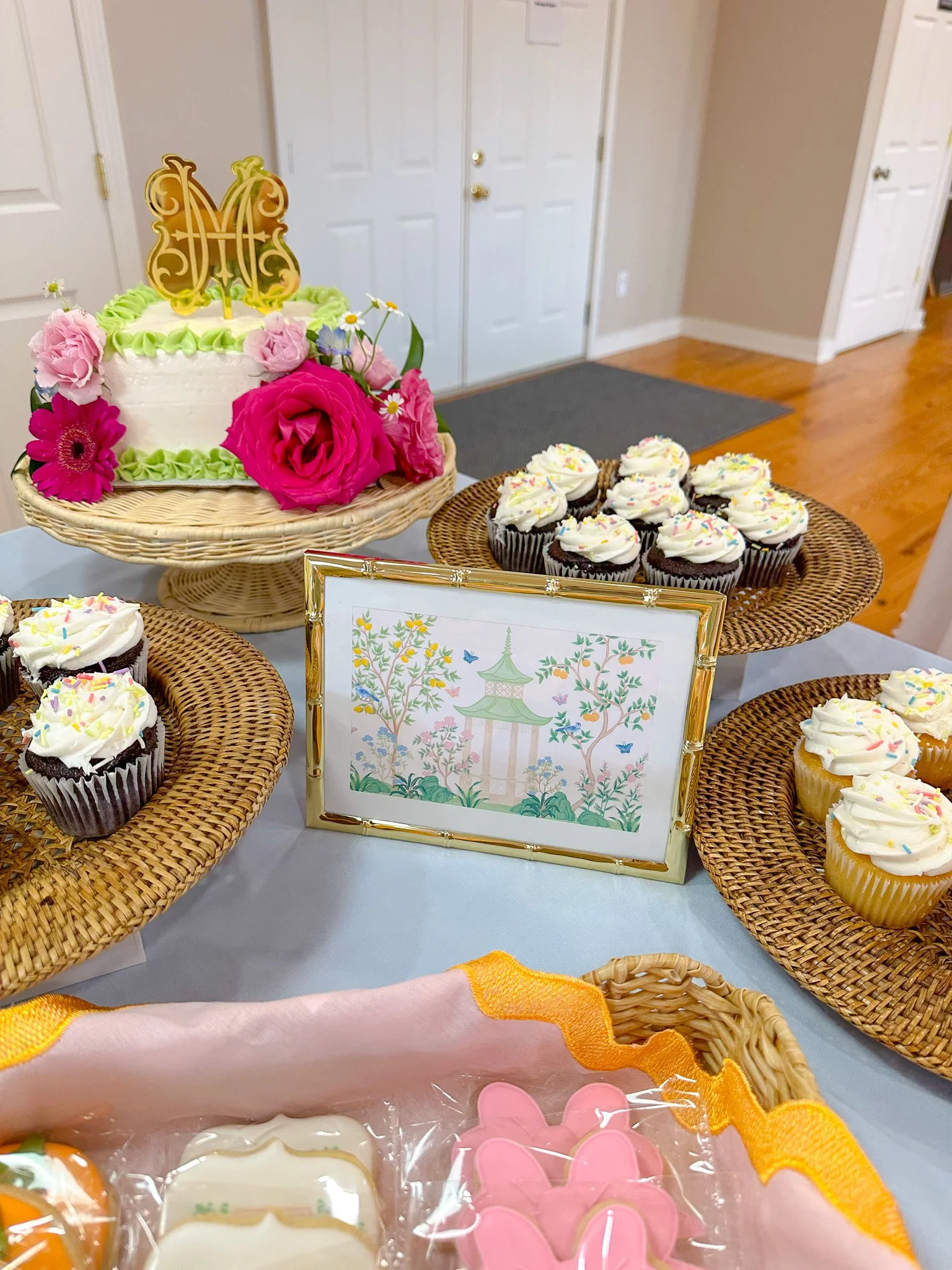 Decorated birthday cake with pink, white, and green frosting, pink roses, and a gold letter topper, along with cupcakes decorated with white frosting and colorful sprinkles, displayed on a table with a framed picture of a pagoda and trees, and small