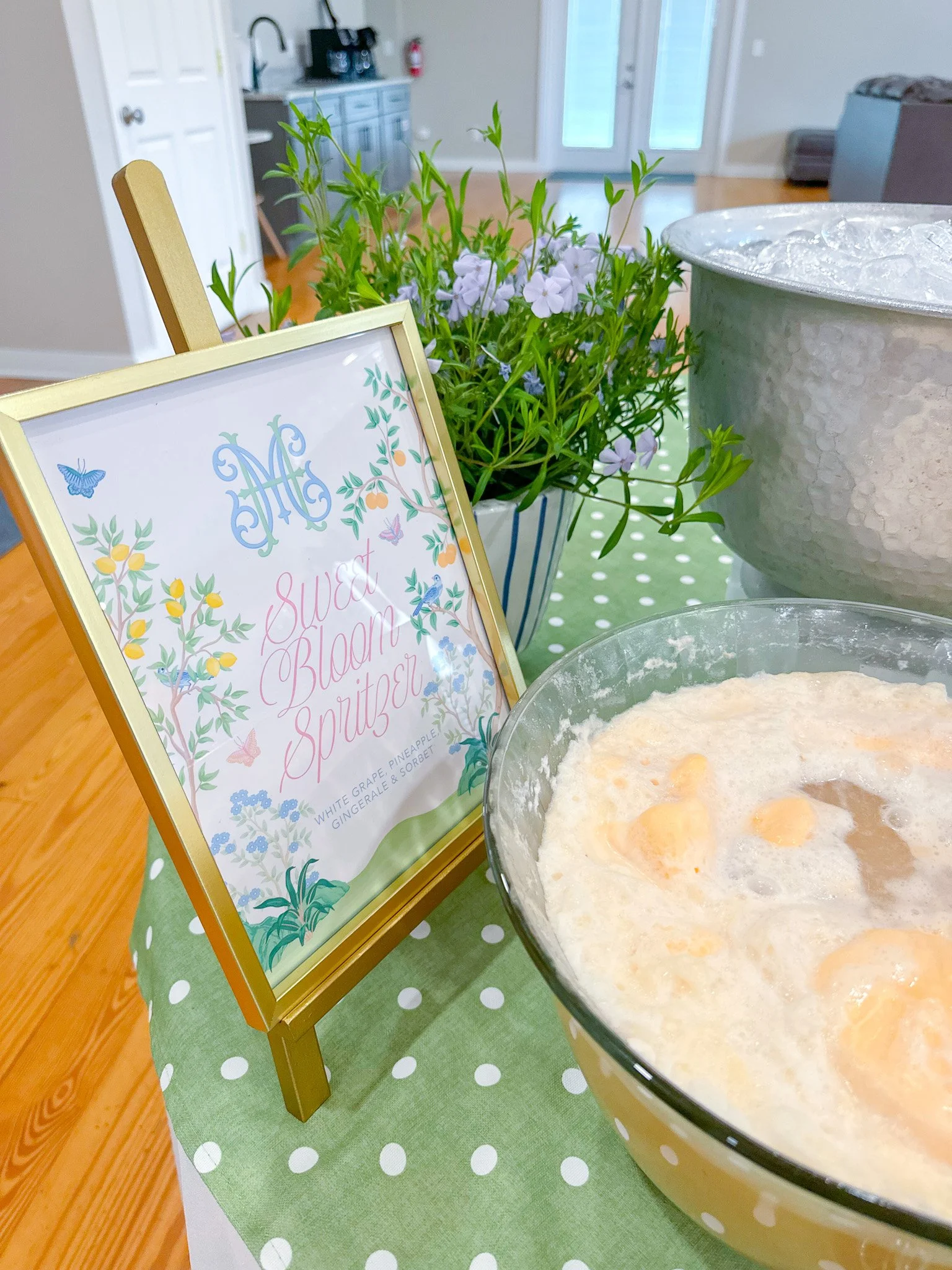 A pitcher of white grape, pineapple, gingerale, and sorbet drink next to a framed sign that reads 'Sweet Bloom Spritzer' and a potted plant with purple flowers, on a green and white polka dot tablecloth.