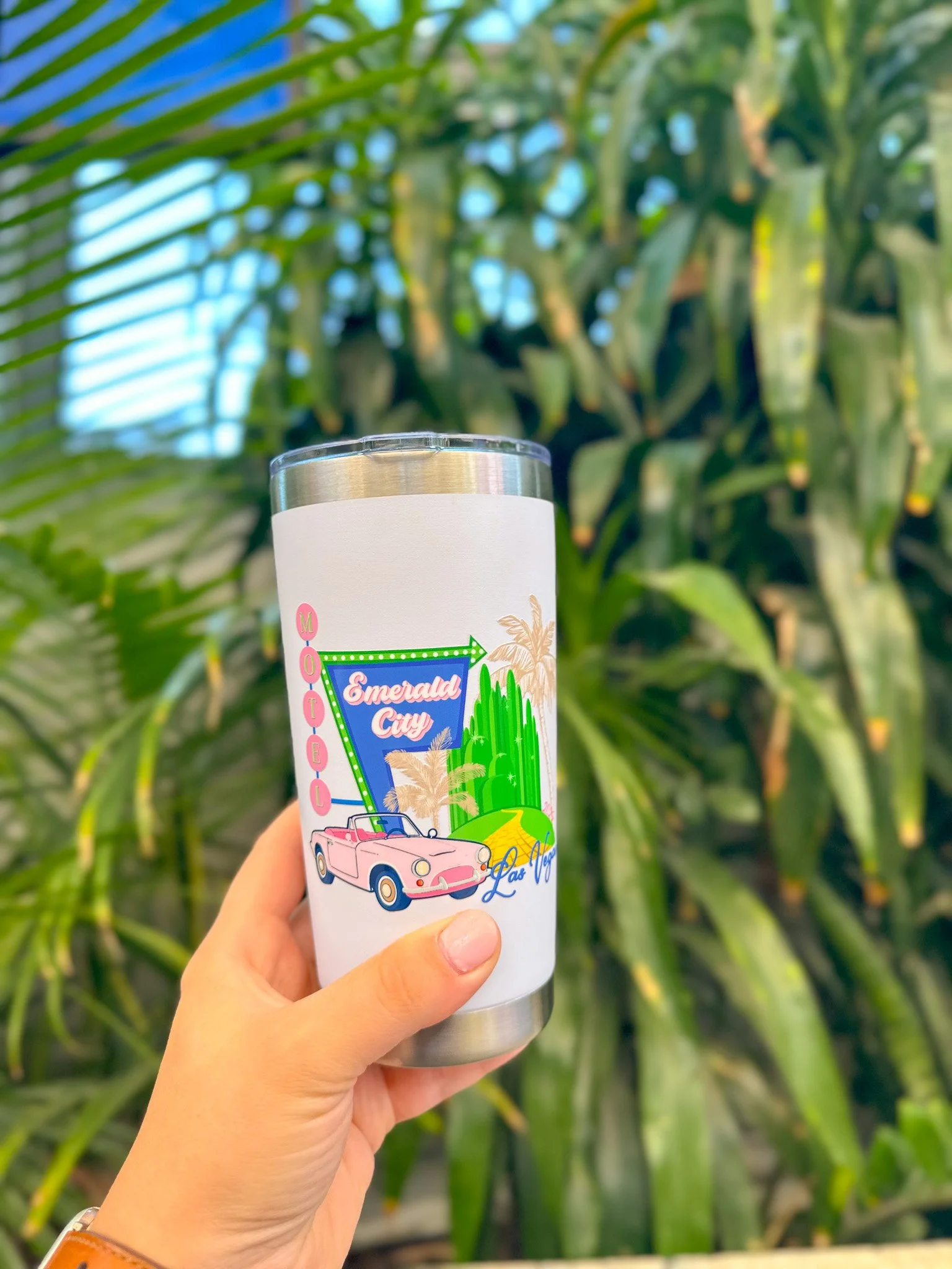 Hand holding a white tumbler with a colorful 'Emerald City Las Vegas' design, featuring a pink convertible car, green cacti, and palm trees, in front of green plants.