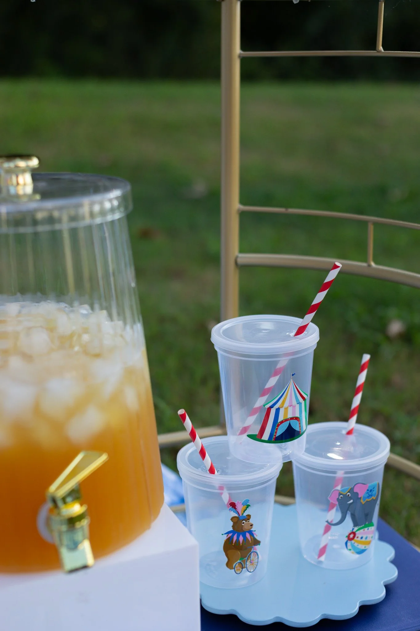 Clear plastic cups with circus-themed stickers, red and white striped straws, a drink dispenser with a spigot, and a gold lid, set outdoors on a white tray with green grass in the background.