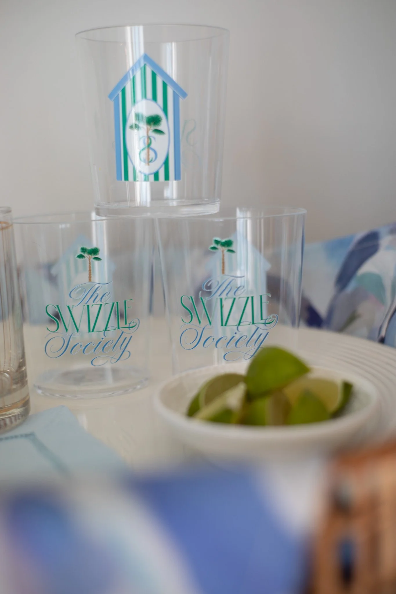 Three clear plastic cups with 'The Swizzle Society' logo, two on the table and one stacked on top, a bowl of lime wedges, and a blurry background.