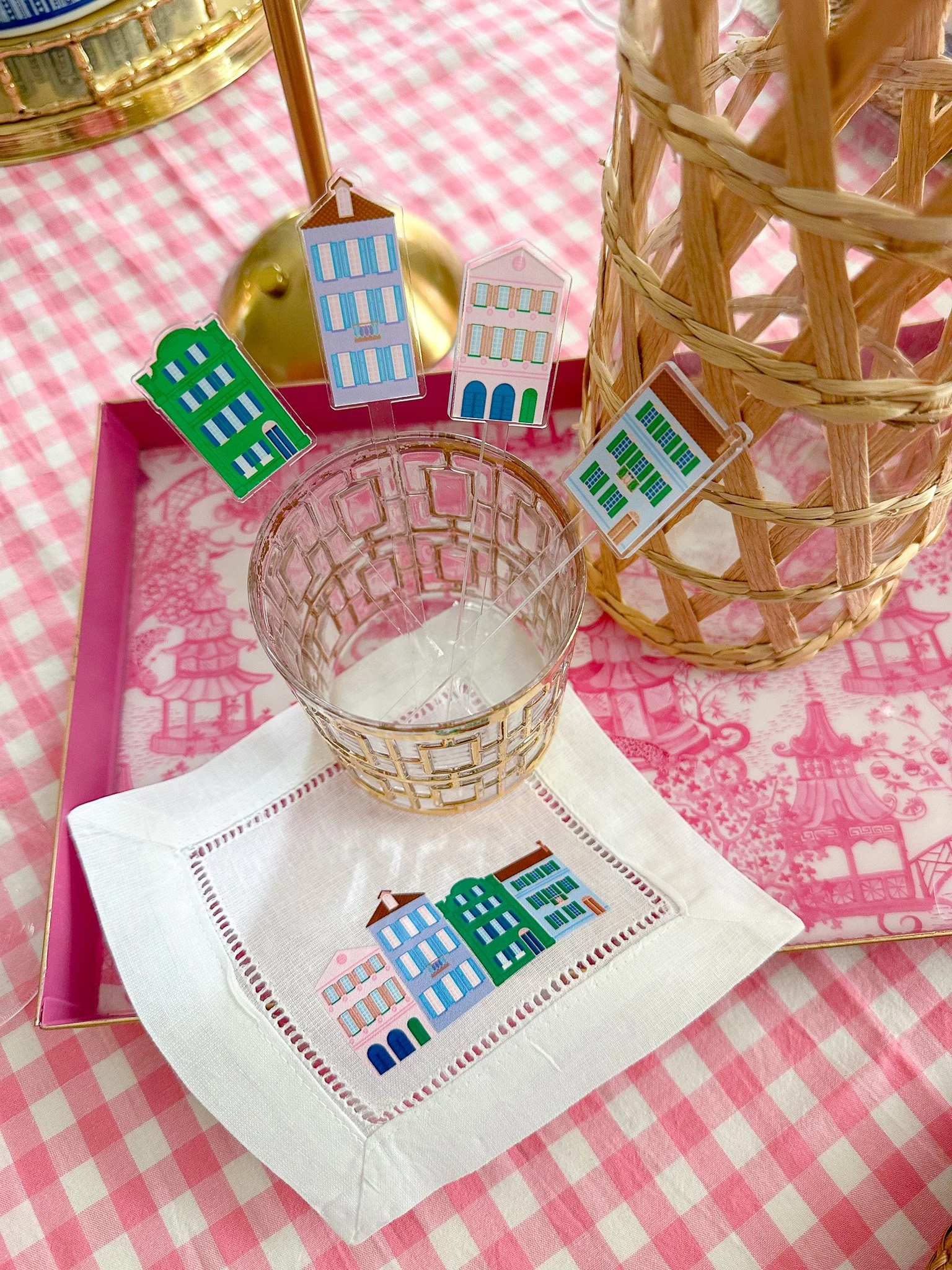 Decorative table setting featuring a glass with paper house pins, a plate of paper house napkins, and a wicker basket on pink checkered tablecloth.