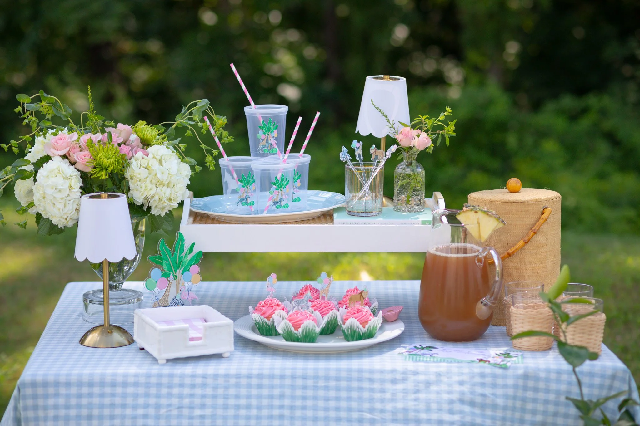 A table set outdoors with pink and white cupcakes, a pitcher of tea with a pineapple slice, beverage cups with pink striped straws, and floral decorations in vases, all on a blue checkered tablecloth.