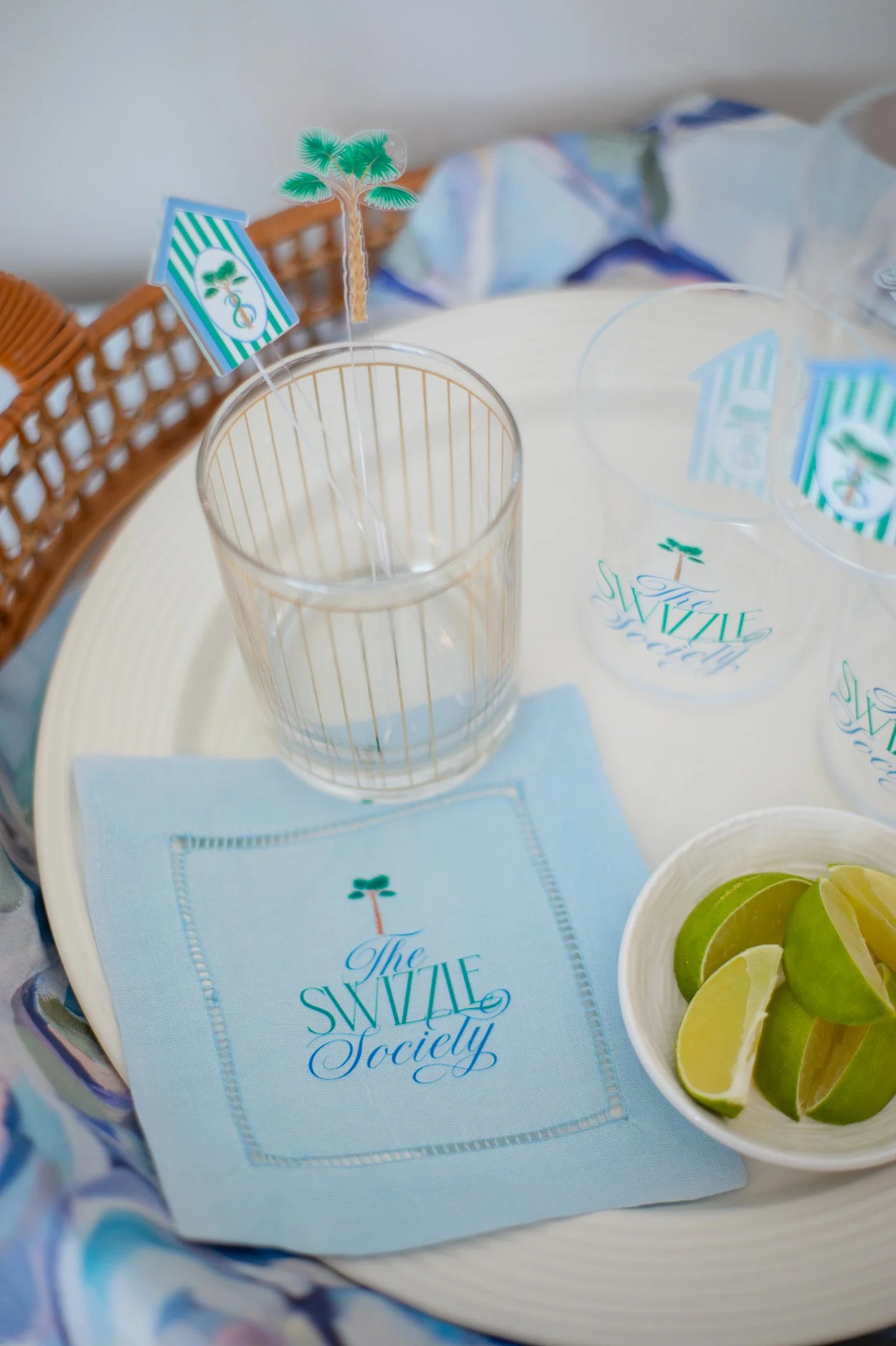 Table setting for The Swizzle Society, including a napkin with the logo, a glass with gold vertical stripes, lime wedges in a dish, and drink stirrers with palm tree and striped flag designs.