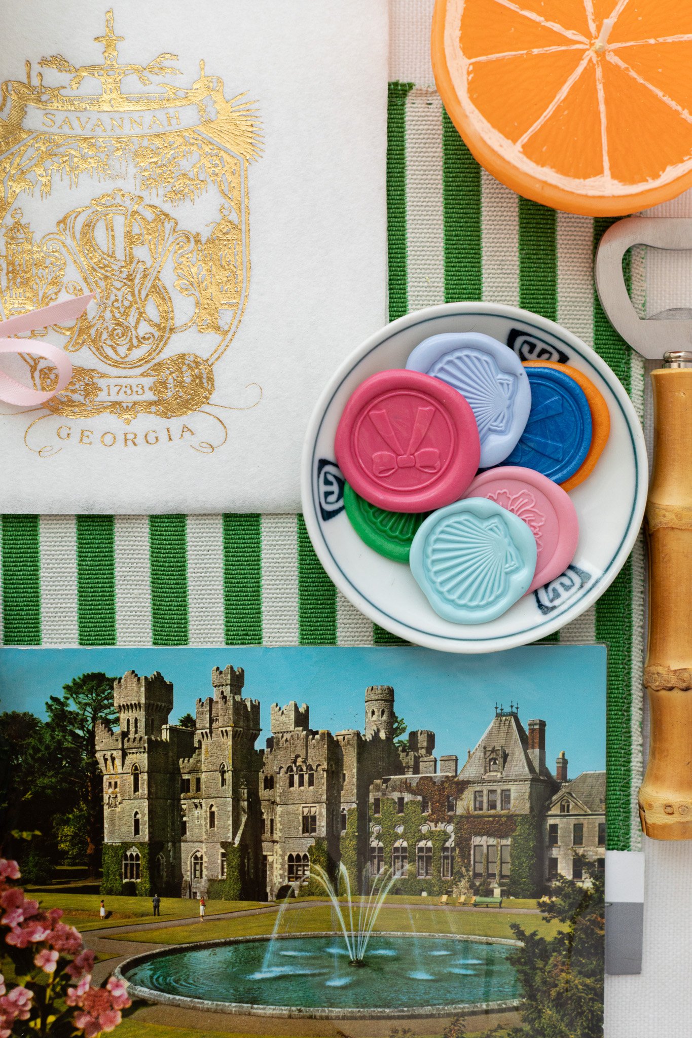 A collection of colorful wax seals, a postcard of a castle with a fountain, and a slice of orange arranged on a green and white striped tablecloth, with a sticker or emblem from Georgia in the top left corner.