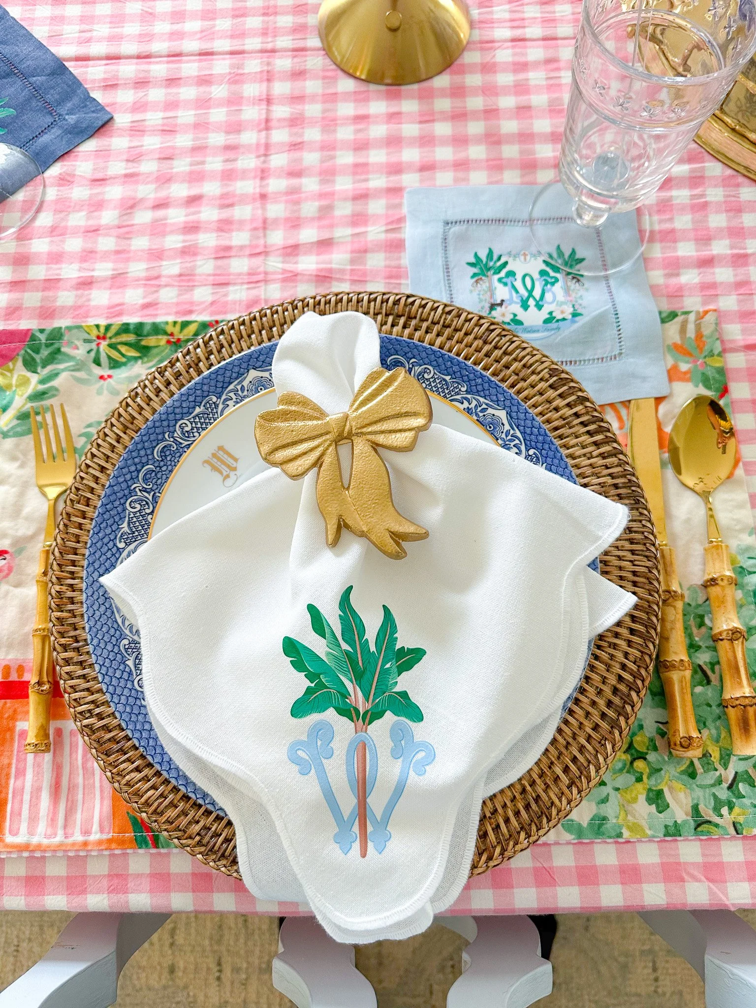 Table setting with decorated napkins, gold utensils, glassware, and a woven placemat on a pink checkered tablecloth, with tropical-themed designs and a gold ornament.