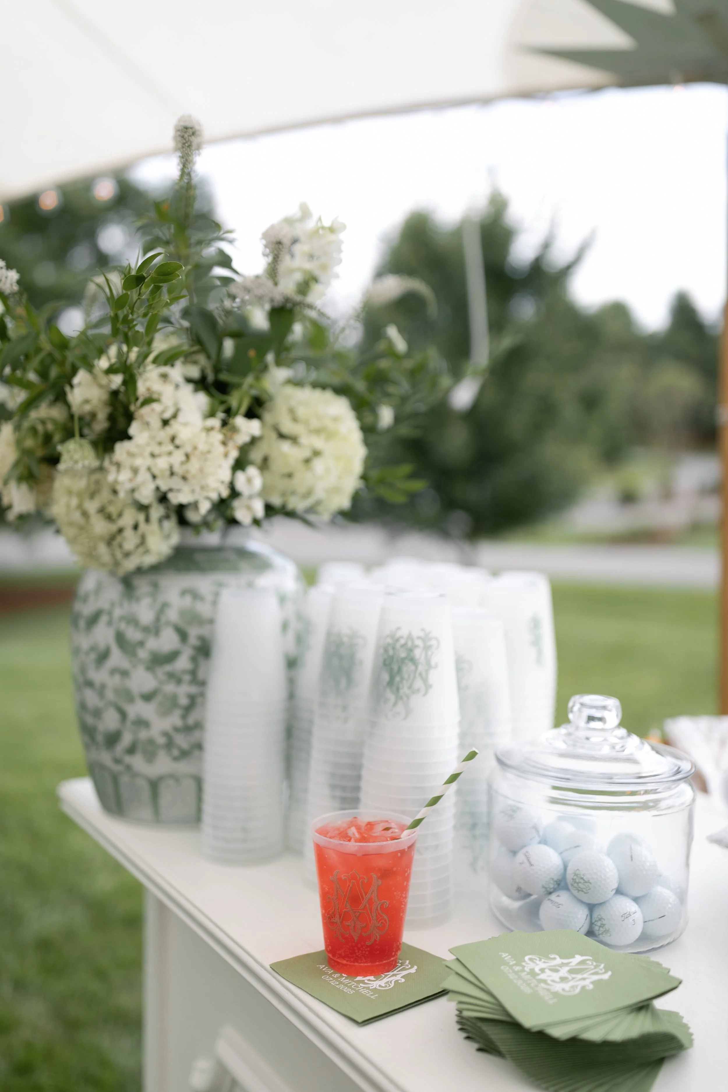 A table with a floral arrangement, stacks of white cups, a jar of golf balls, and a pink drink with a striped straw outdoors.