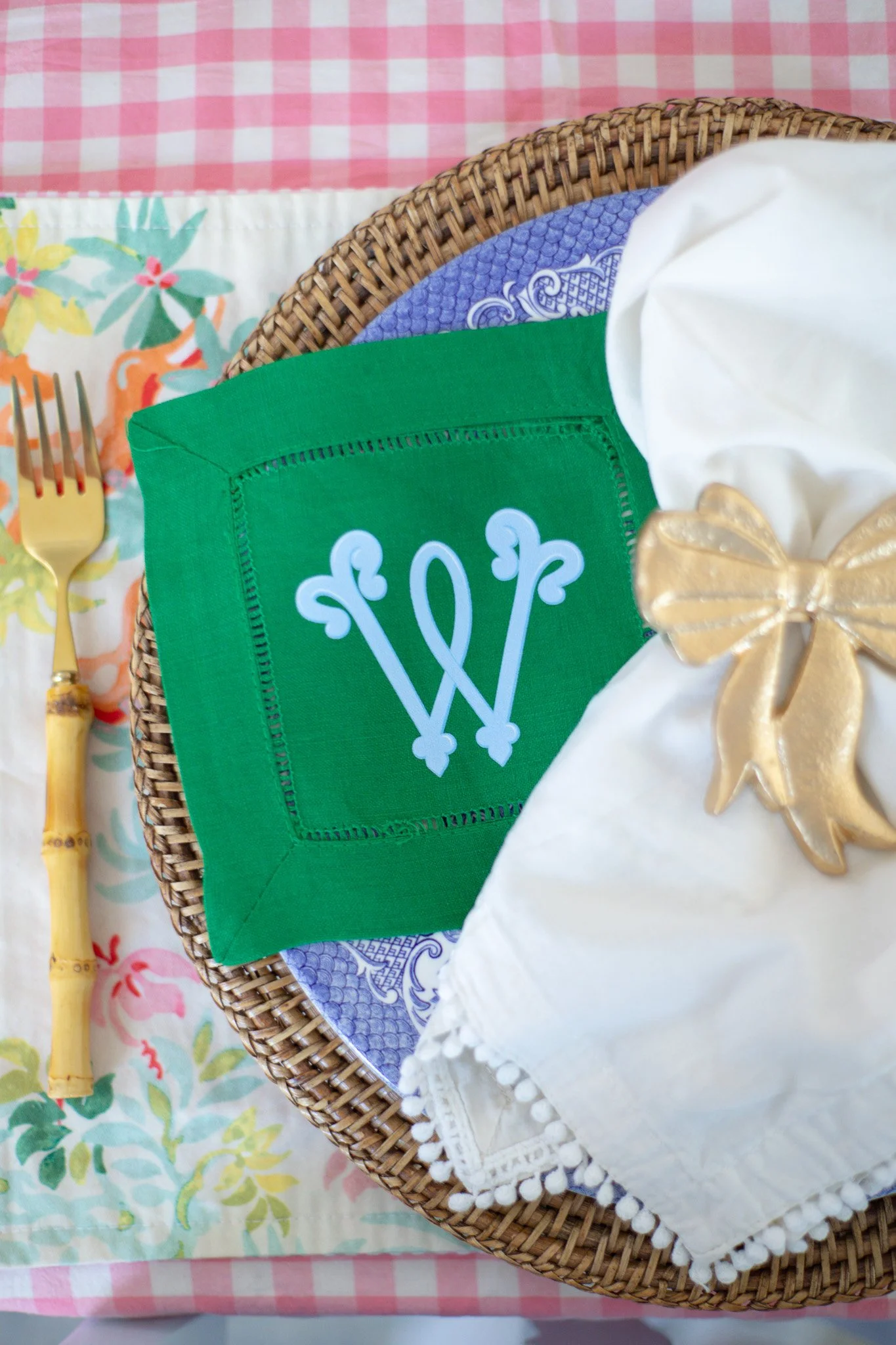 Table setting featuring a green cloth napkin with a white decorative monogram, a gold fork with a bamboo handle, a white cloth napkin with a gold bow, and a woven placemat, all on a pink and white checkered tablecloth.