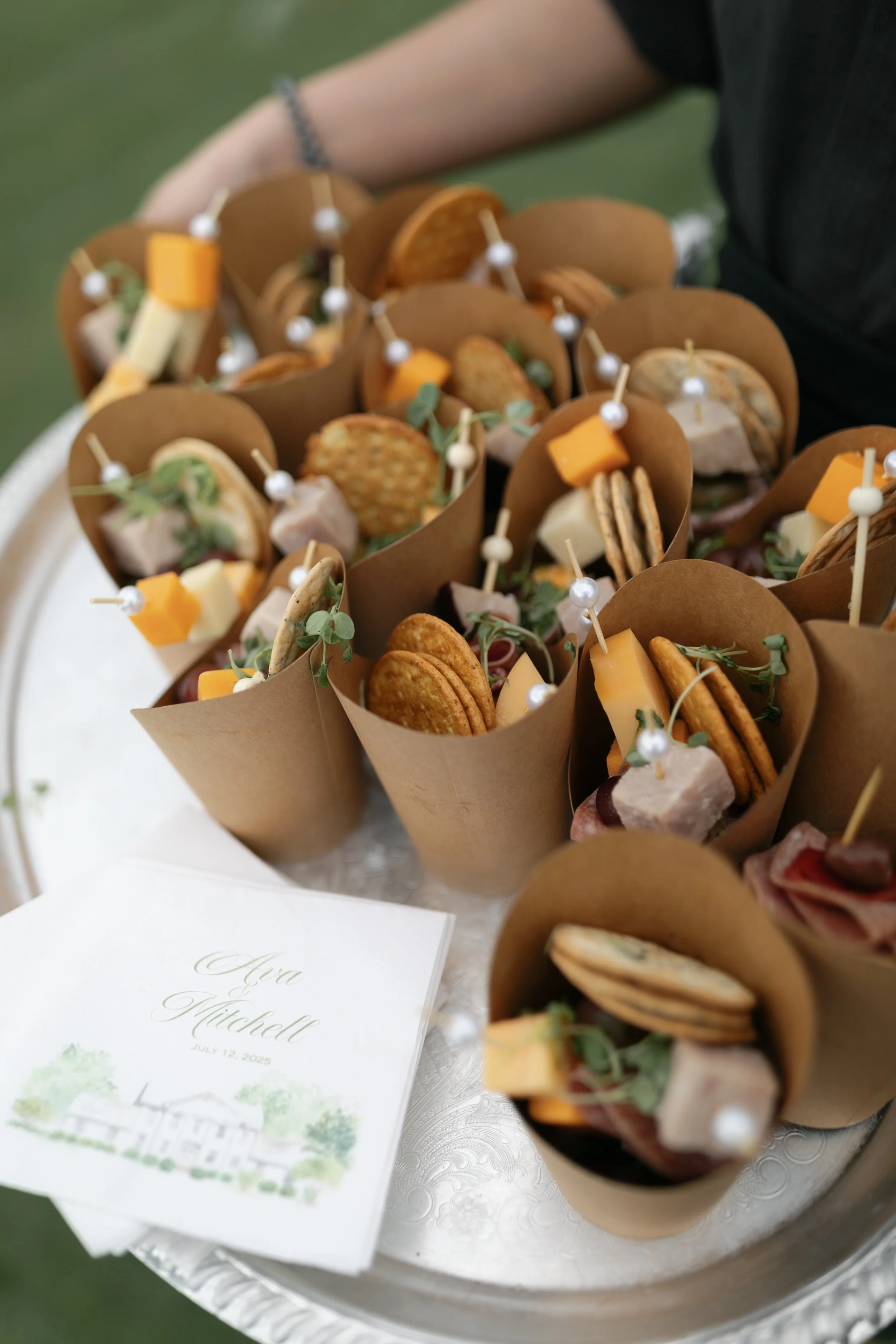 Tray of mini sandwiches with cheese, crackers, meats, and garnishes, arranged in brown paper cones at a formal event.