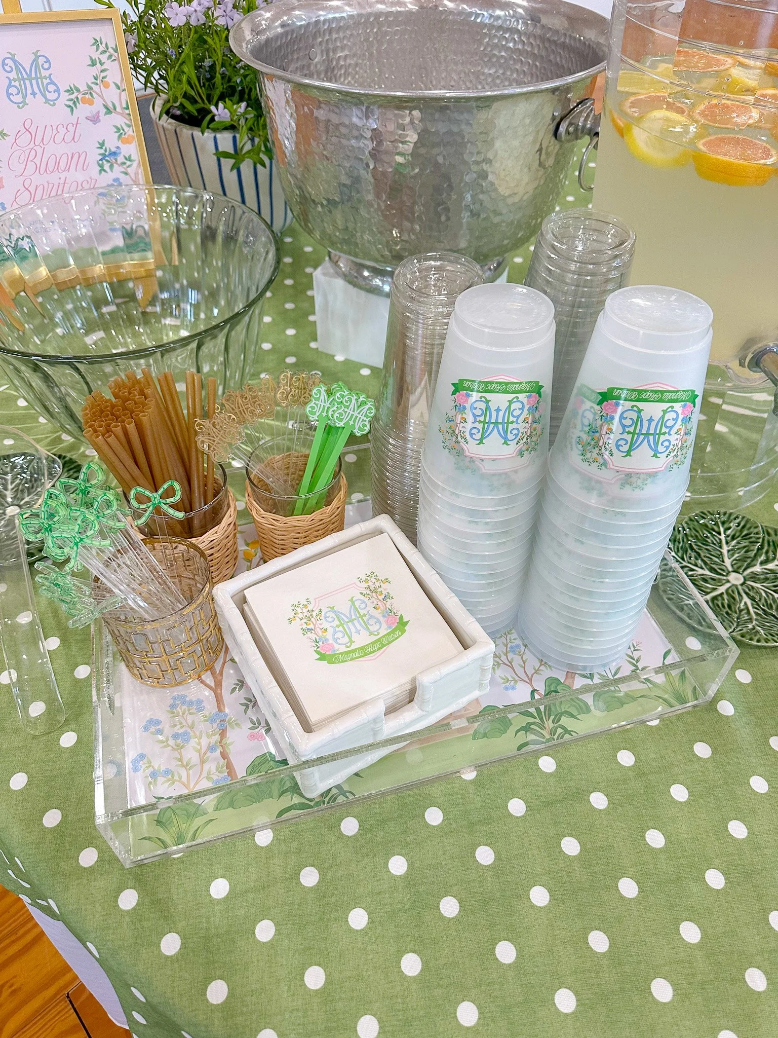 A beverage station with cups labeled 'Magnolia Home Queen', a punch bowl filled with lemon-lime soda with lemon slices, a large ice bucket, and various straws and drink stirrers on a polka-dotted green tablecloth.